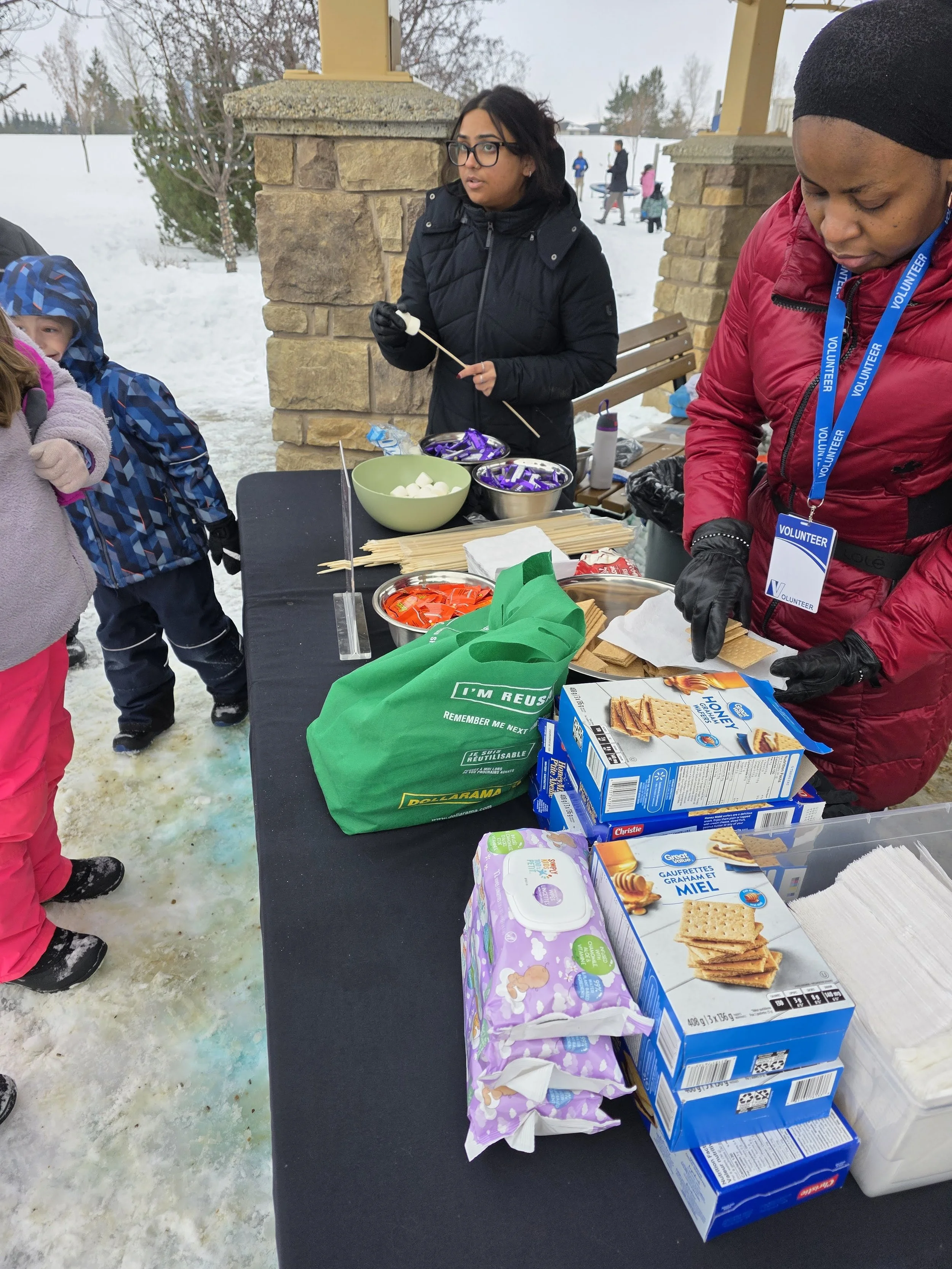 People preparing and serving snacks outdoors at a winter event, with snow on the ground, a table with boxes of graham crackers and marshmallows, and children waiting nearby.