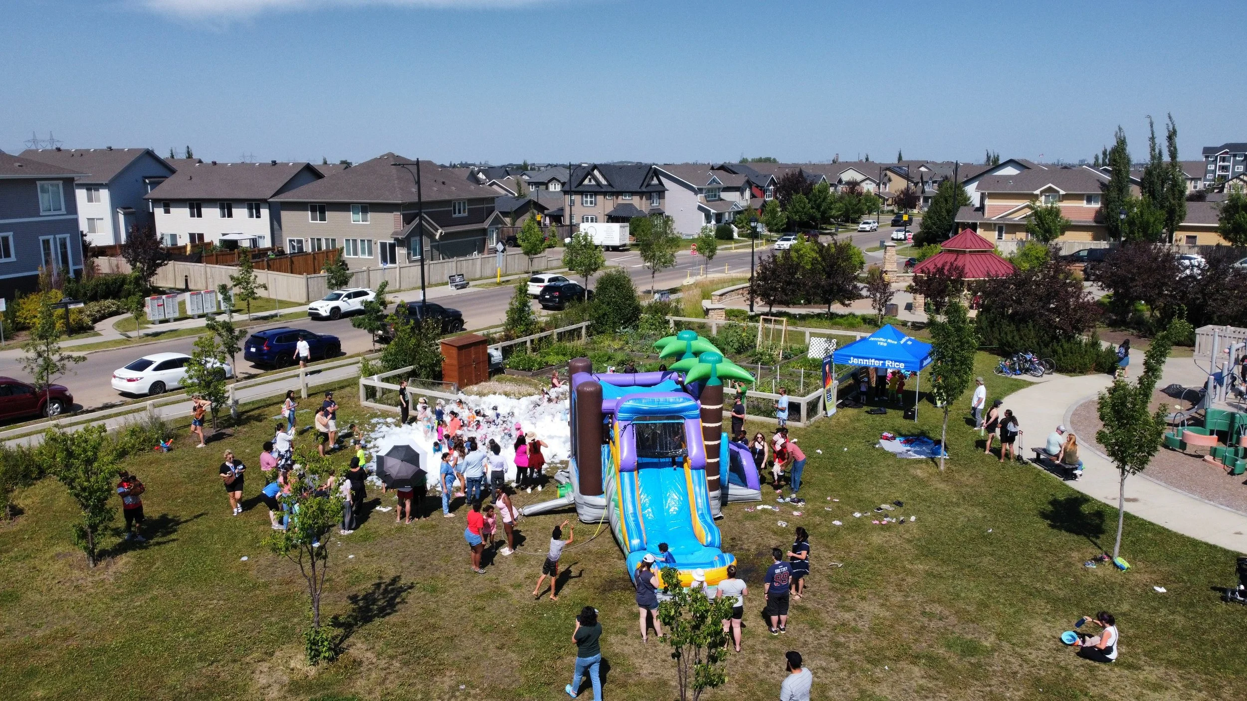 A community outdoor party with children and adults around a large inflatable water slide, foam, and trees in a residential neighborhood park.