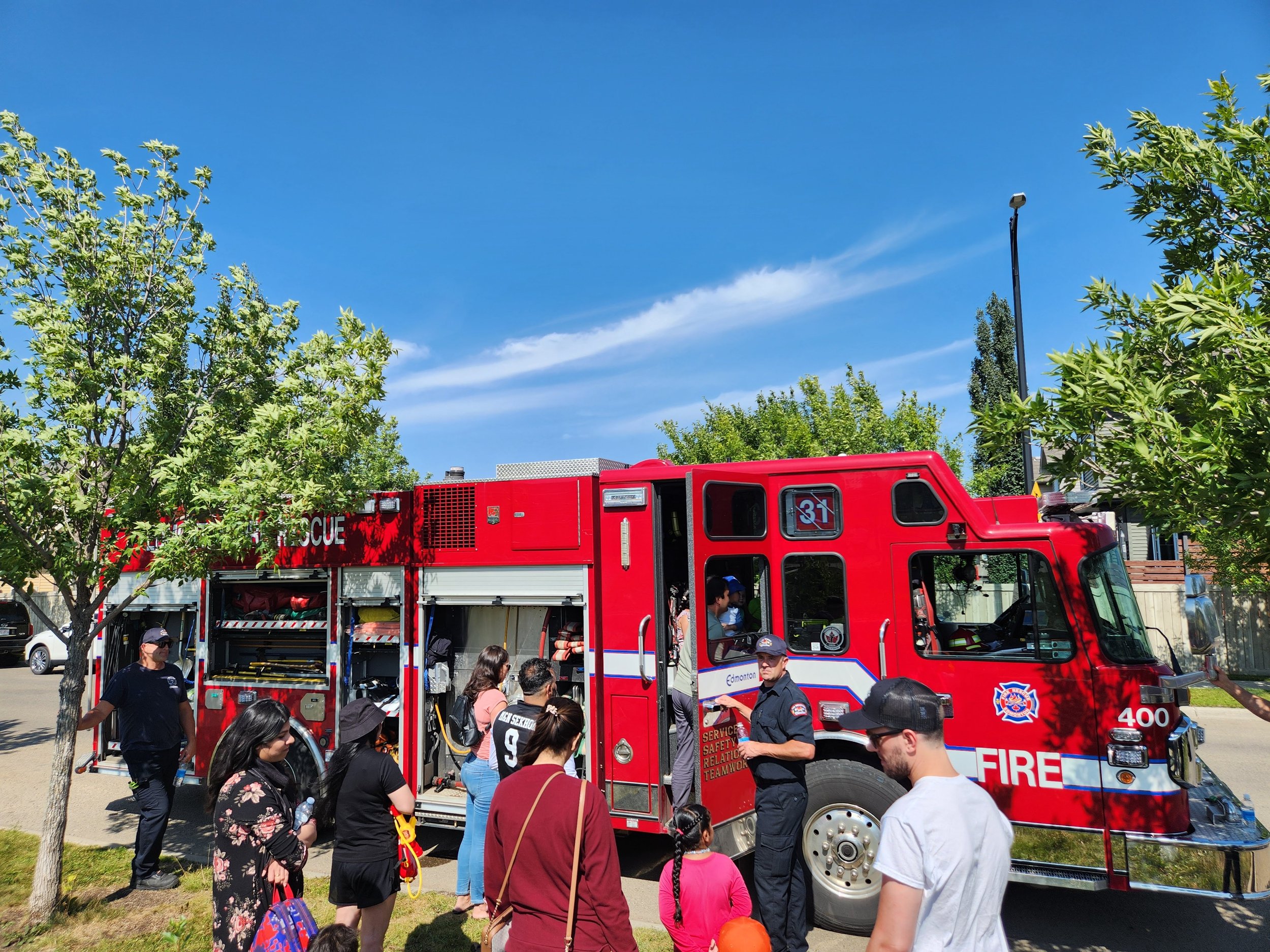People gather around a red fire truck parked on a street with trees and a blue sky.