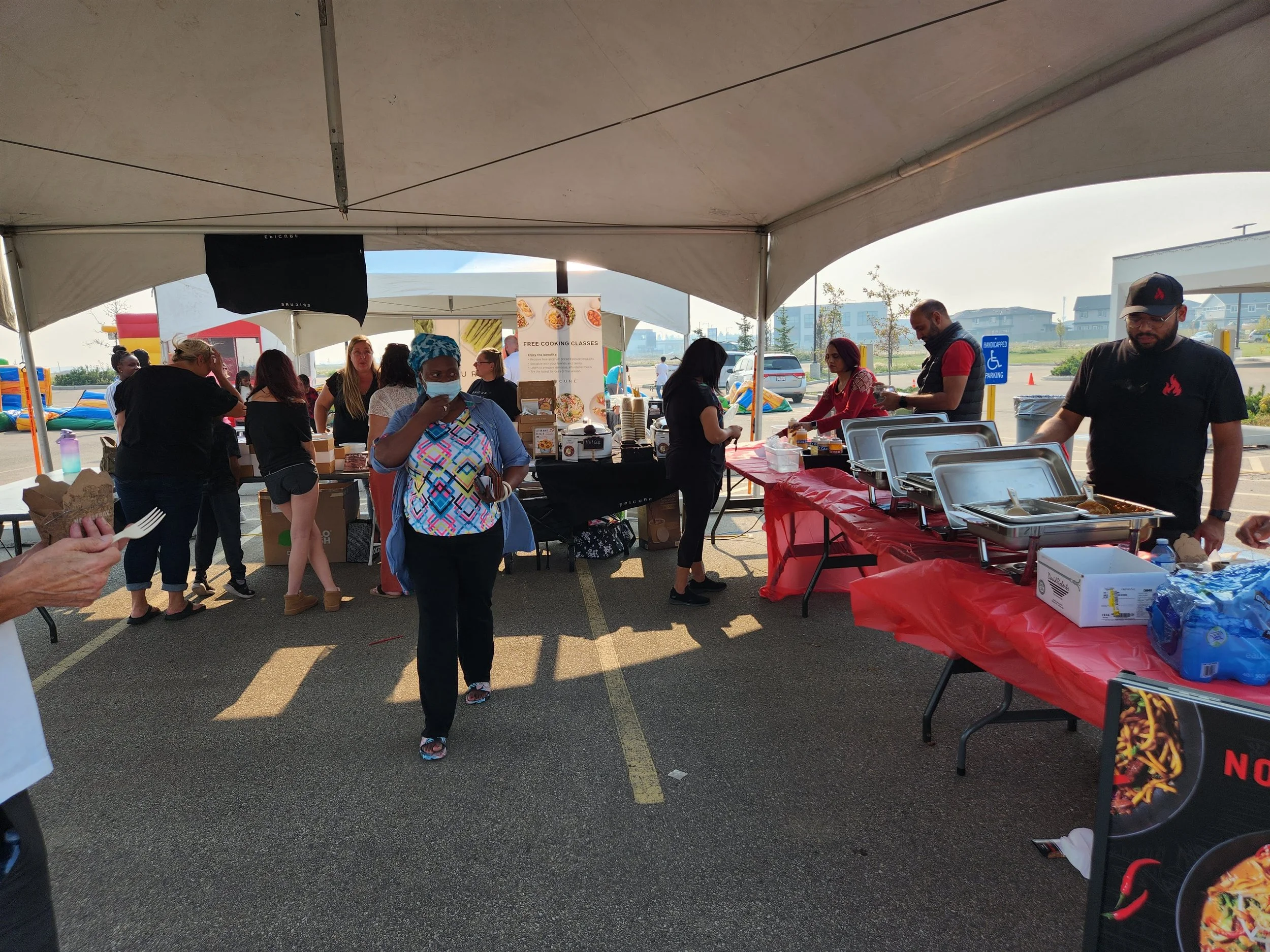 People attending a food stall under a large tent at an outdoor event. Some are serving and others waiting or eating. Various food items and catering tables are visible, with a few people wearing masks.