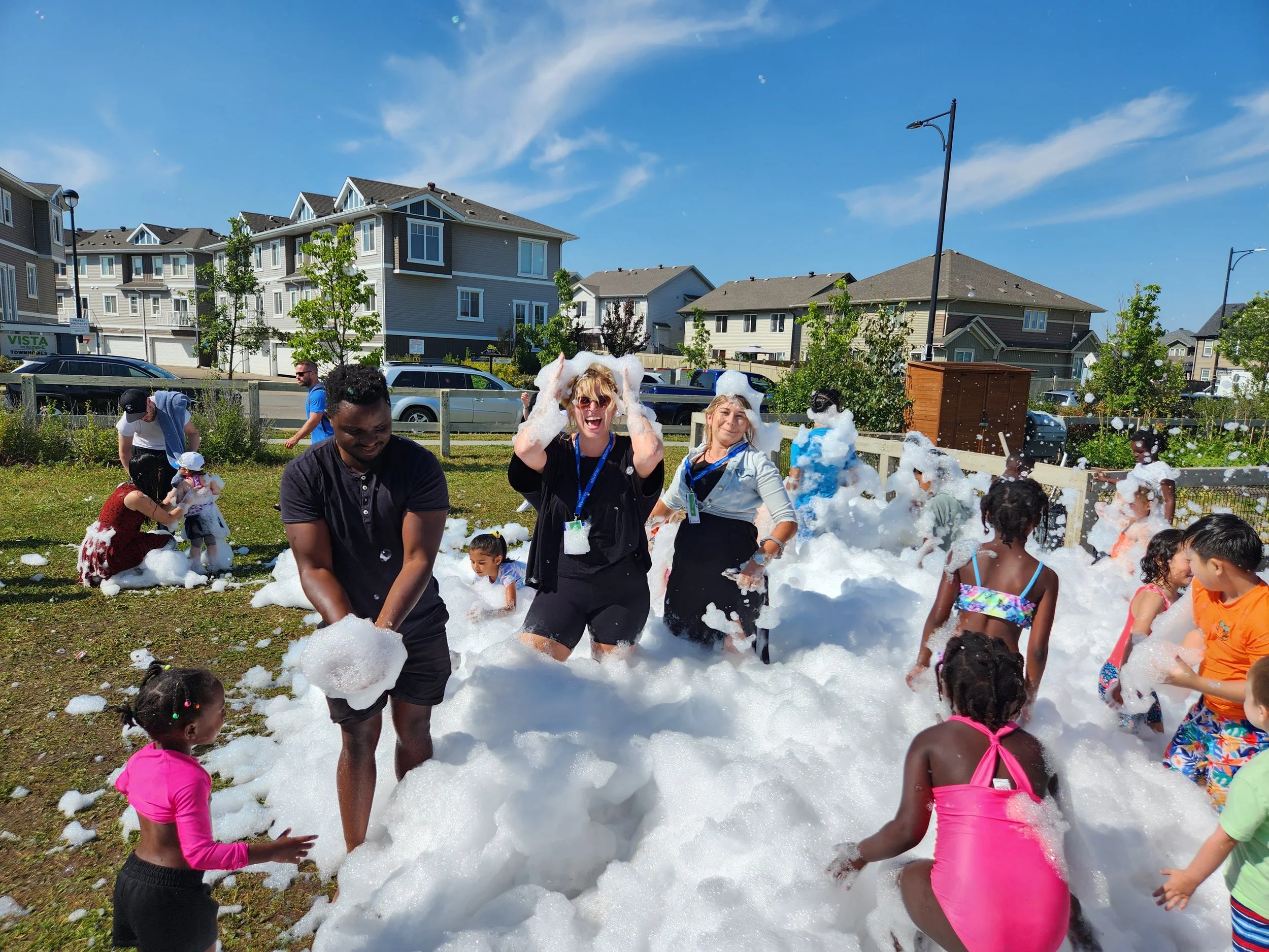Children and adults playing in a foam party outdoors on a sunny day, with foam covering the grass and people laughing and enjoying.