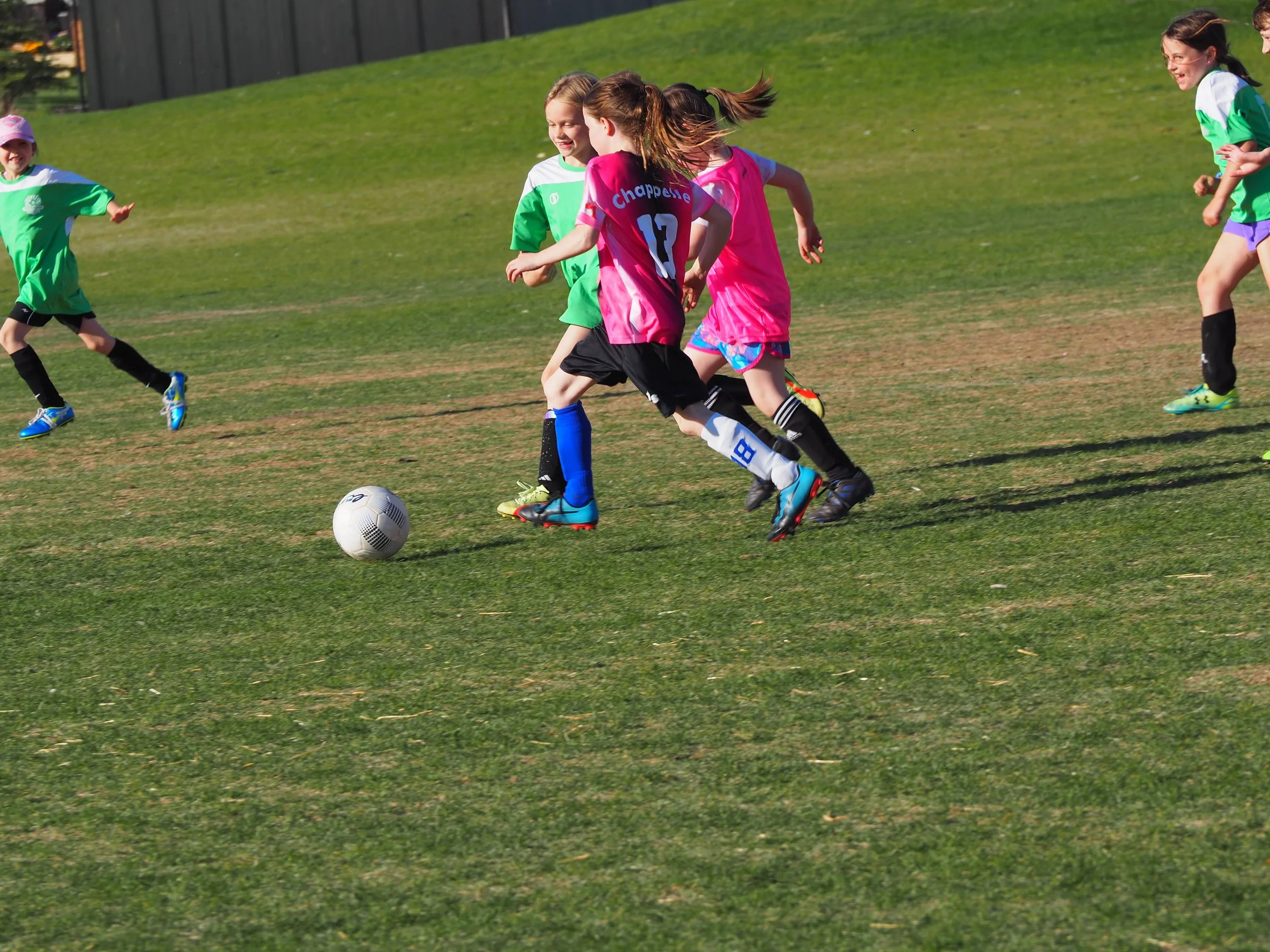 Girls playing soccer on a grassy field with some running towards the ball, wearing colorful sports uniforms and cleats.