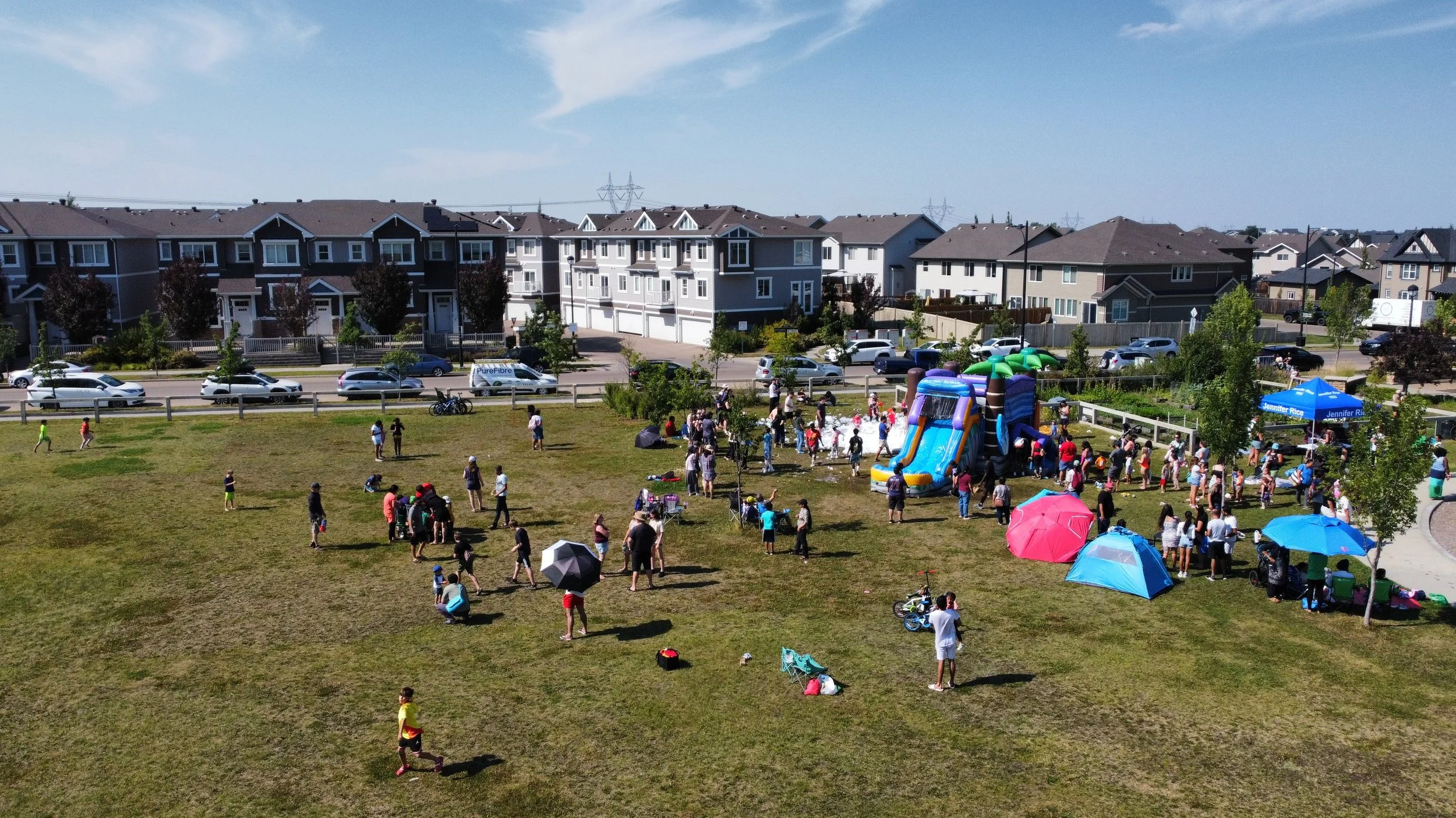 A community park filled with children and adults enjoying a day outdoors. There are colorful umbrellas, tents, and a large inflatable water slide. Families are gathered around, some sitting on the grass, others participating in activities, on a sunny