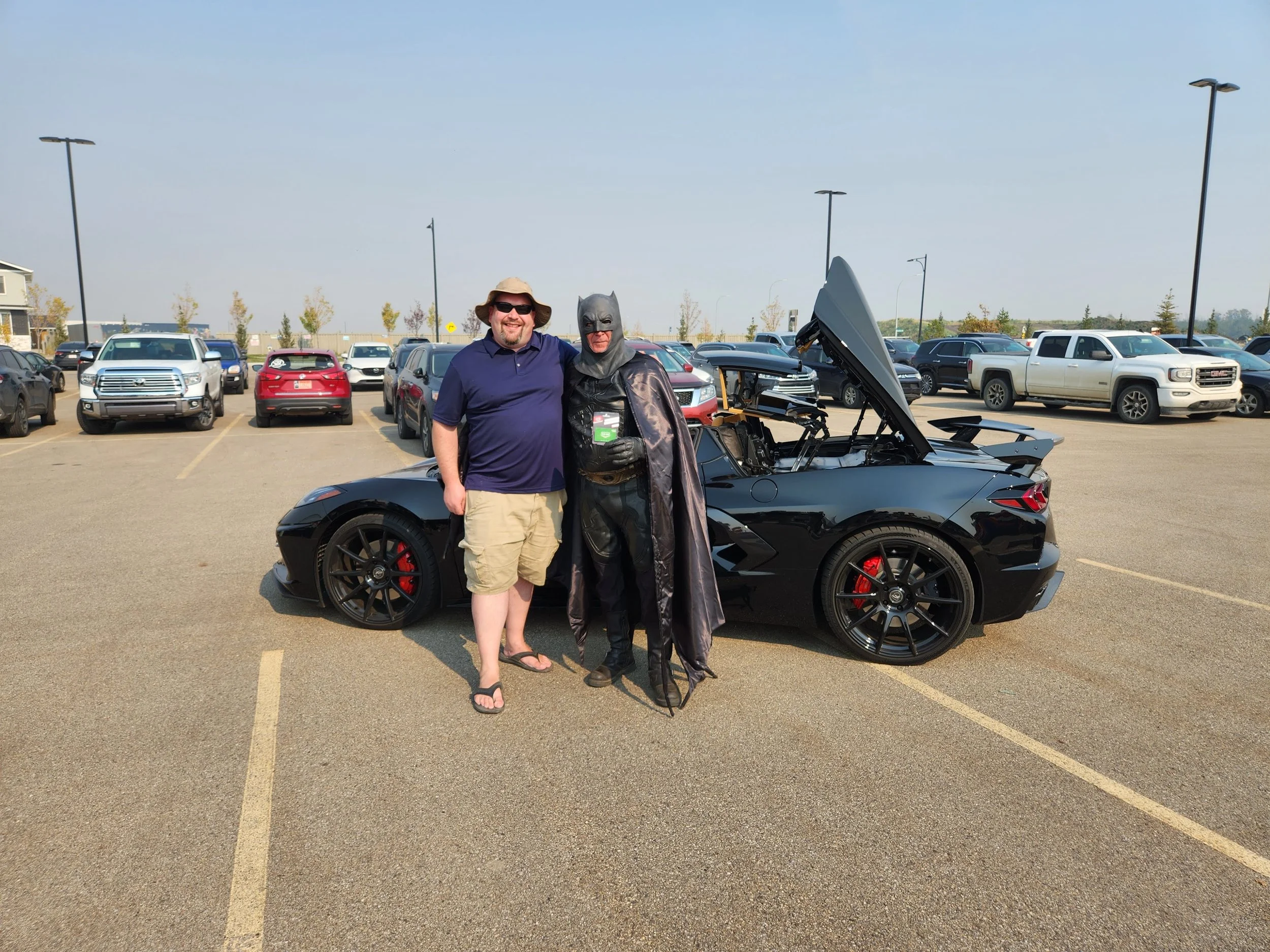 Two men stand next to a black sports car in a parking lot. One man is casually dressed in a navy polo, shorts, flip-flops, and a hat, smiling. The other man is dressed as Batman, with a cape and mask. The car has its top down, and the hood is open.