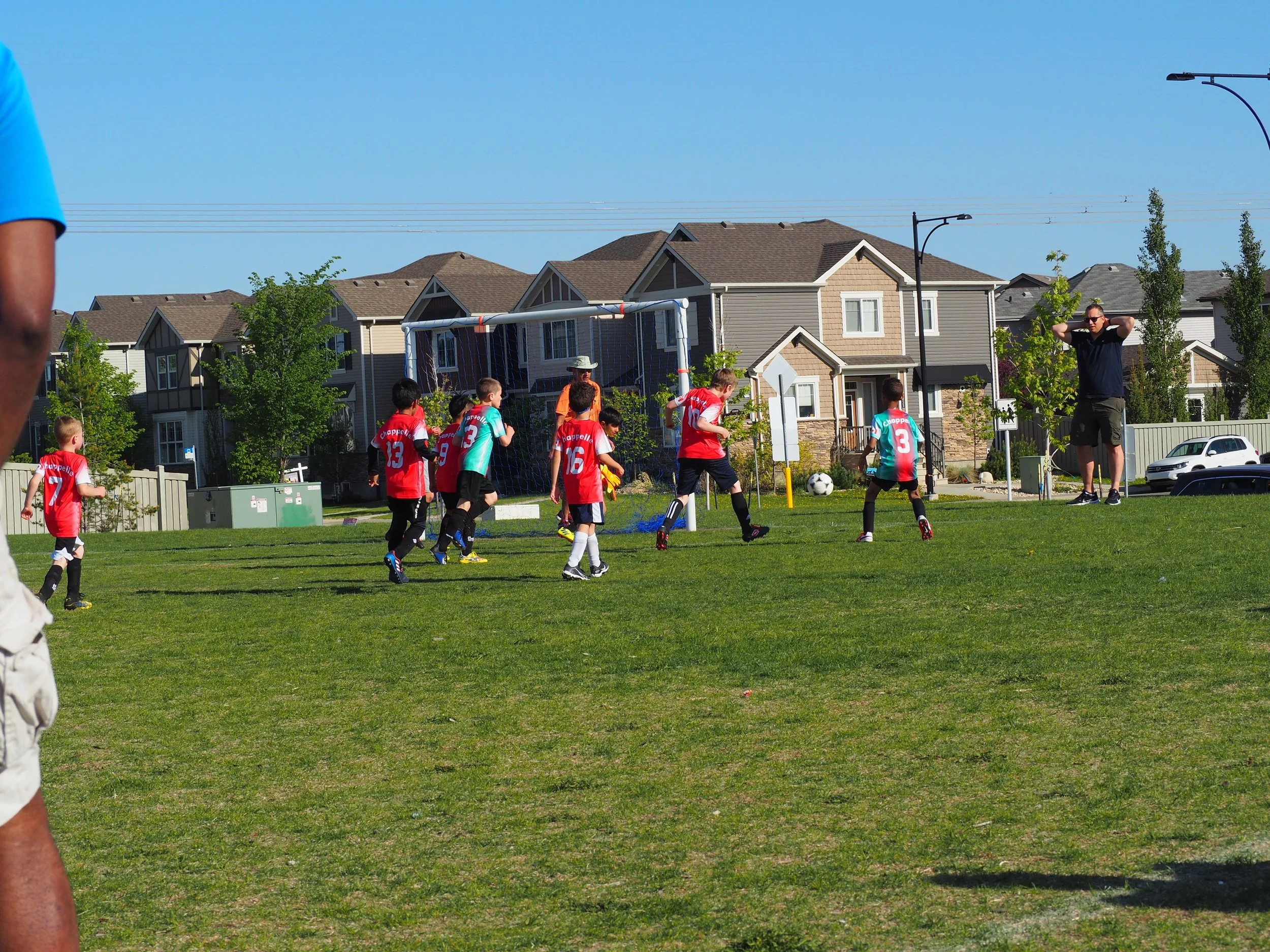 Kids playing soccer on a grassy field in a residential neighborhood, with a coach supervising, houses in the background, and a clear blue sky.