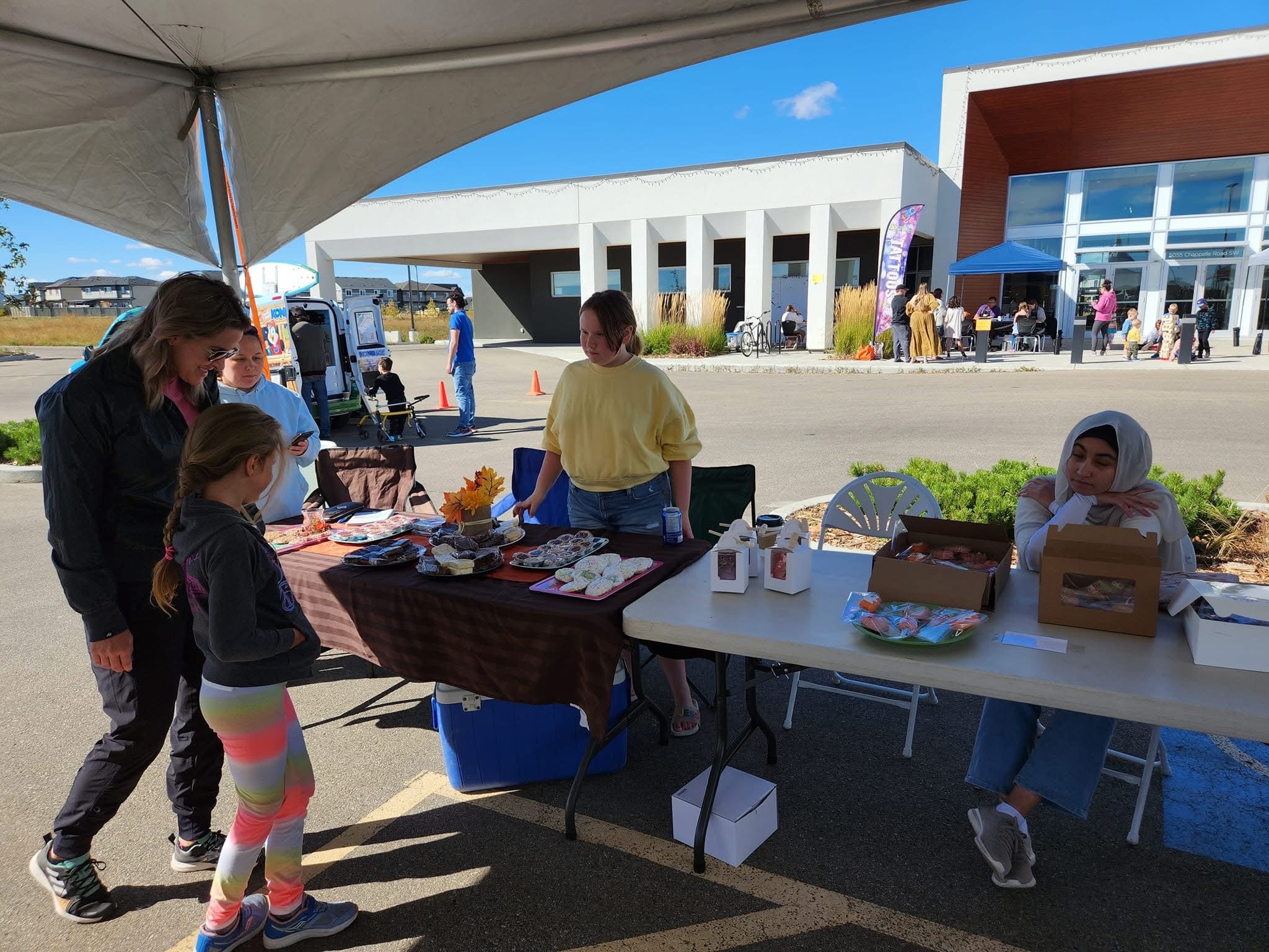 People at an outdoor market stall with baked goods under a canopy on a sunny day, with a modern building in the background.