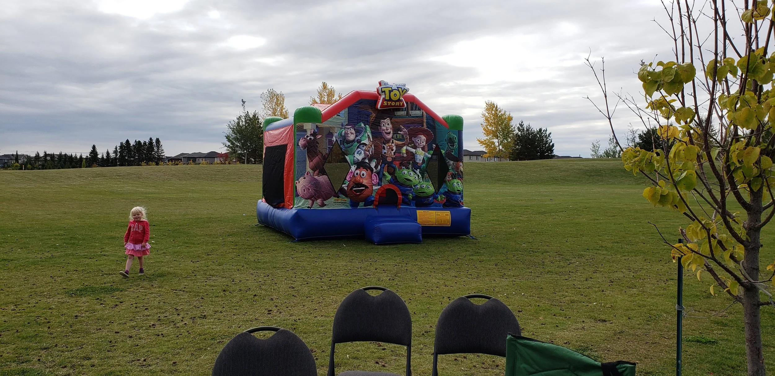 Colorful inflatable bounce house with Toy Story characters on a grassy field, with a child in a pink dress playing nearby and chairs in the foreground, under a cloudy sky.