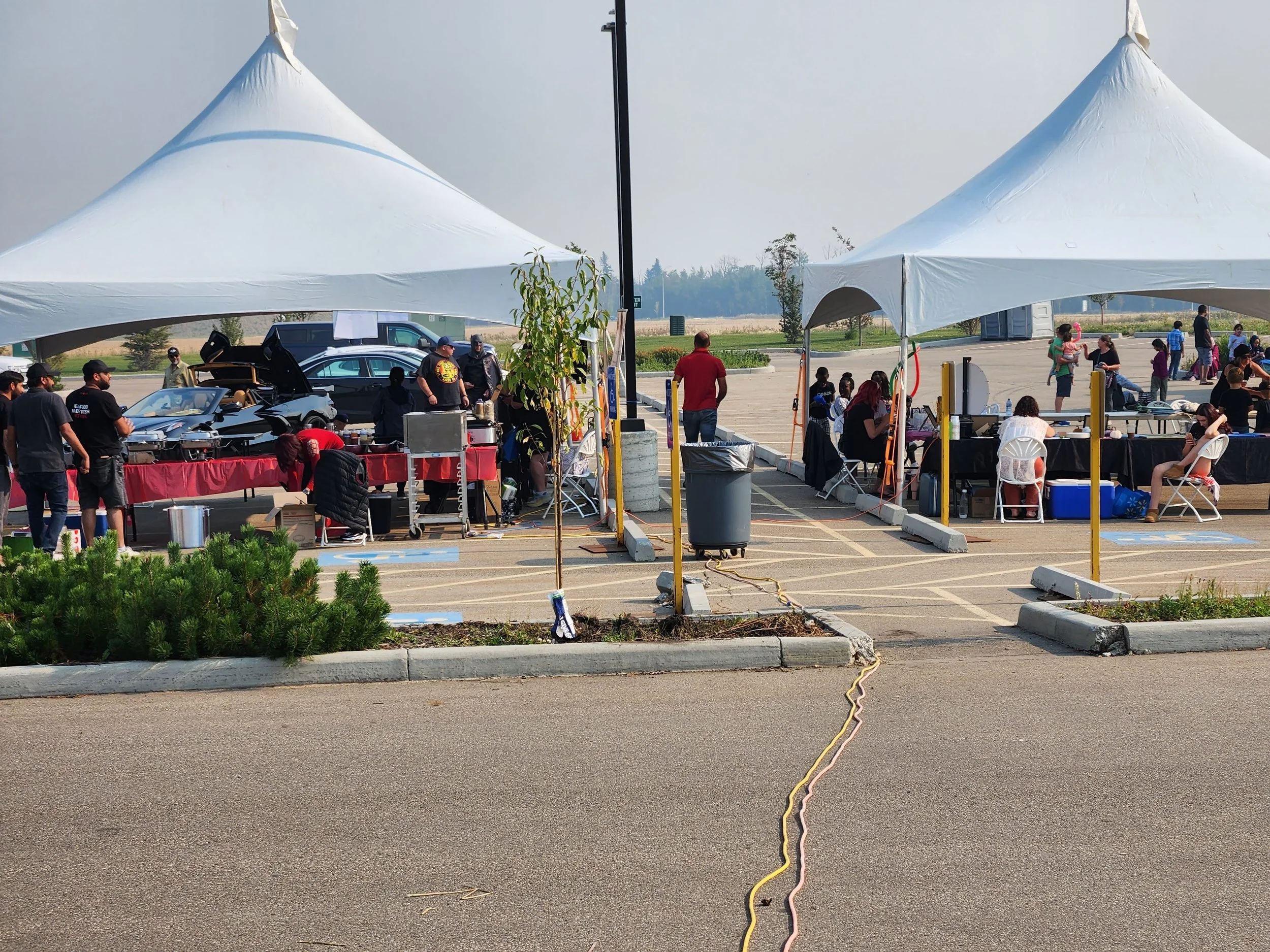 Outdoor car show with two large white tents set up in a parking lot. Under the tents, people are gathered around cars, tables, and chairs, with some standing and some sitting. Vehicles are parked nearby, and there are people in the background walking