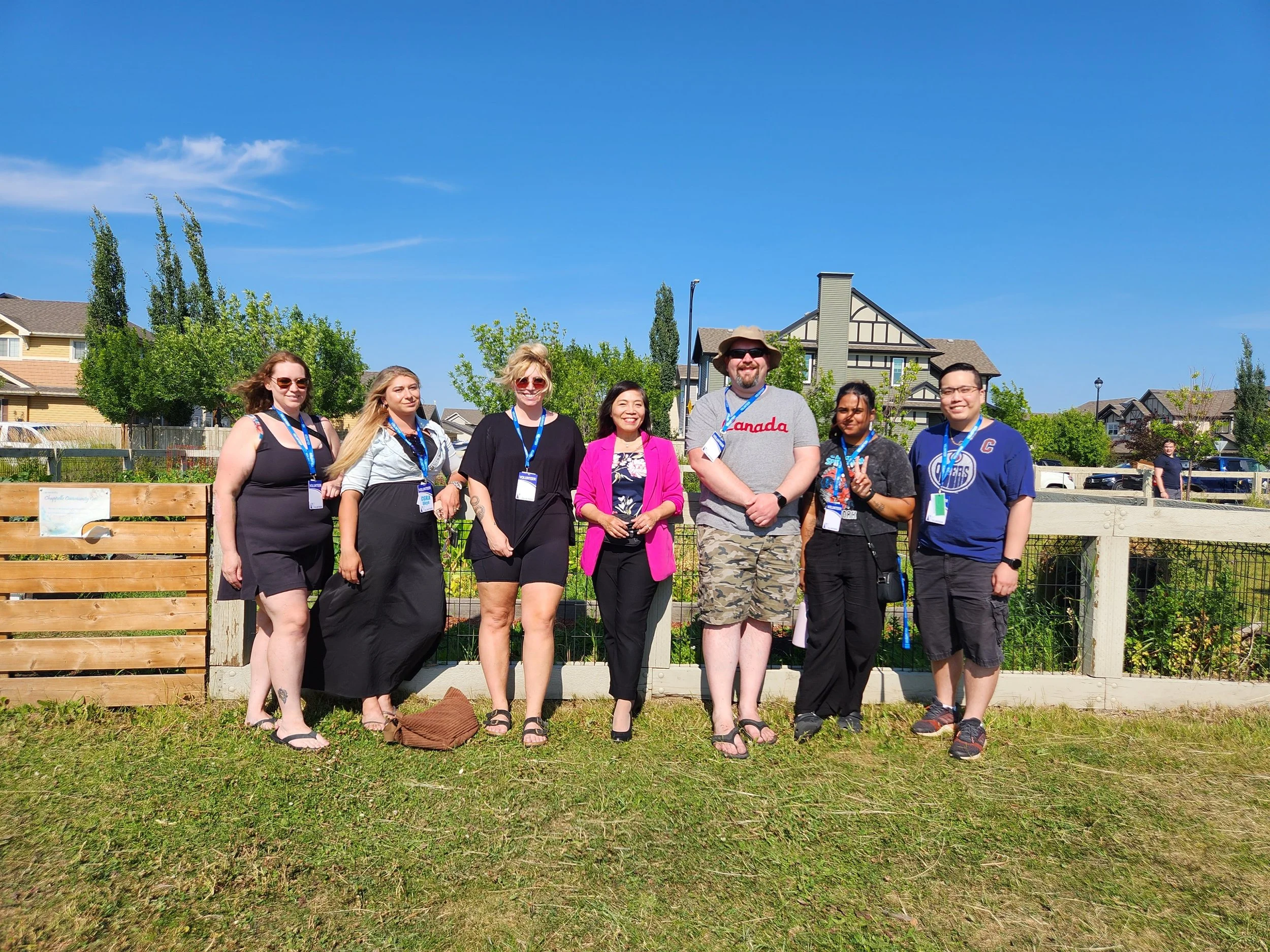 Group of eight diverse people standing outdoors in front of a wooden fence, smiling for a photo on a sunny day with clear blue skies, residential houses, and trees in the background.