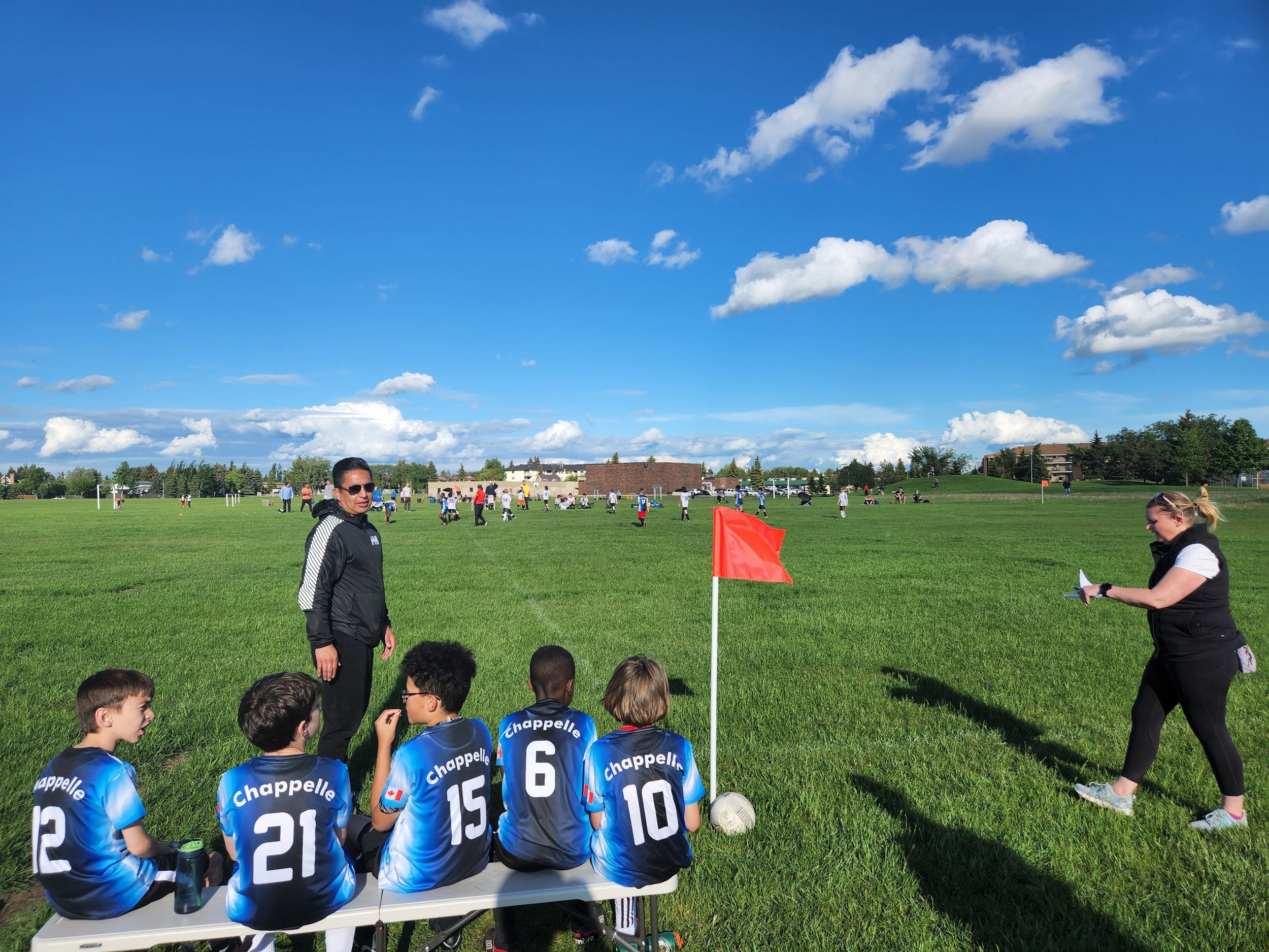 Juvenile soccer players in blue jerseys sitting on a bench on the sideline of a soccer field, with a coach talking to one and a woman taking notes nearby, under a blue sky with scattered clouds.