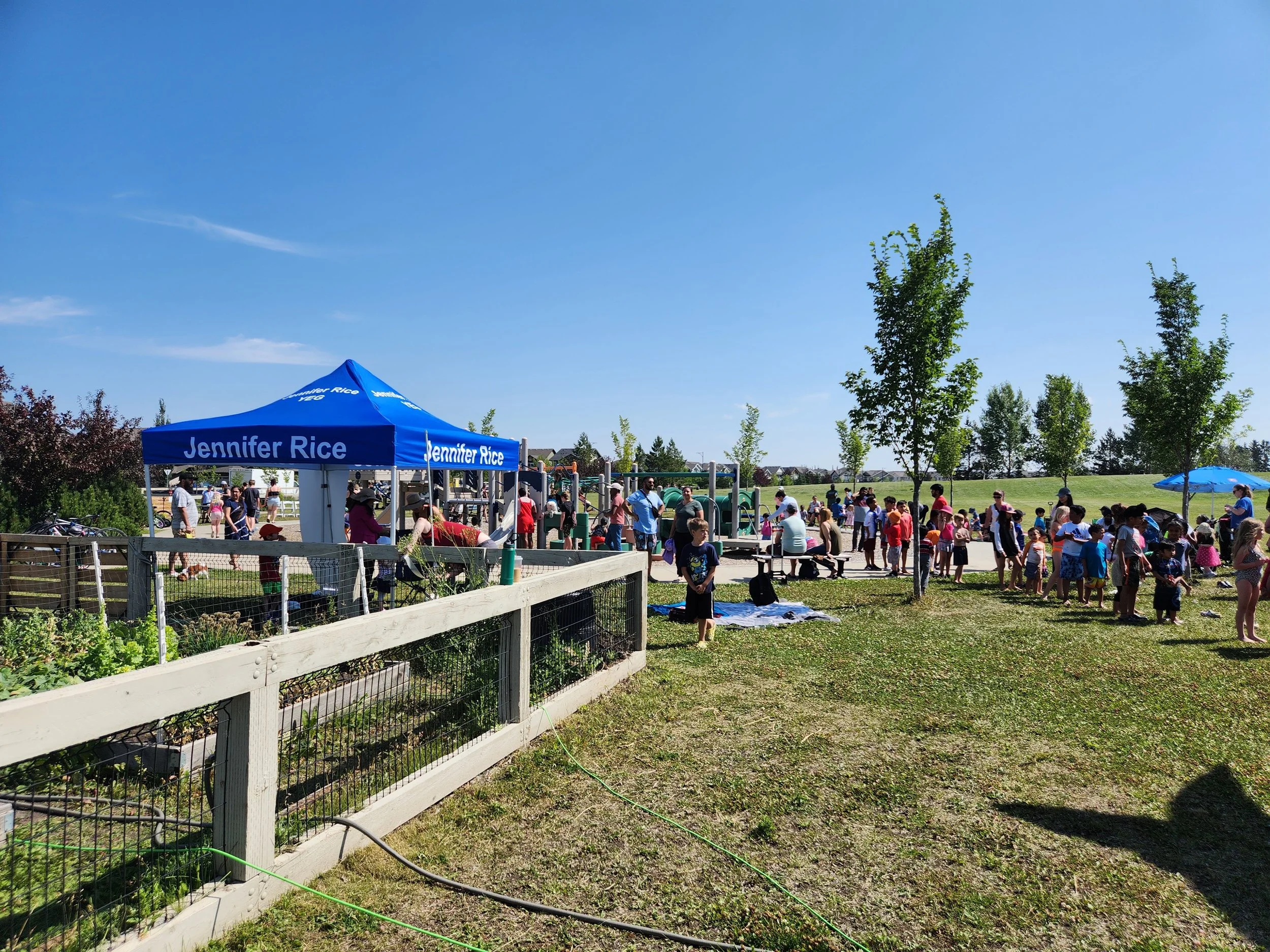 A large outdoor gathering with many children and adults in a park on a sunny day. There is a blue canopy tent with 'Jennifer Rice' written on it, and people are playing, talking, and walking around near playground equipment and open grassy areas, wit