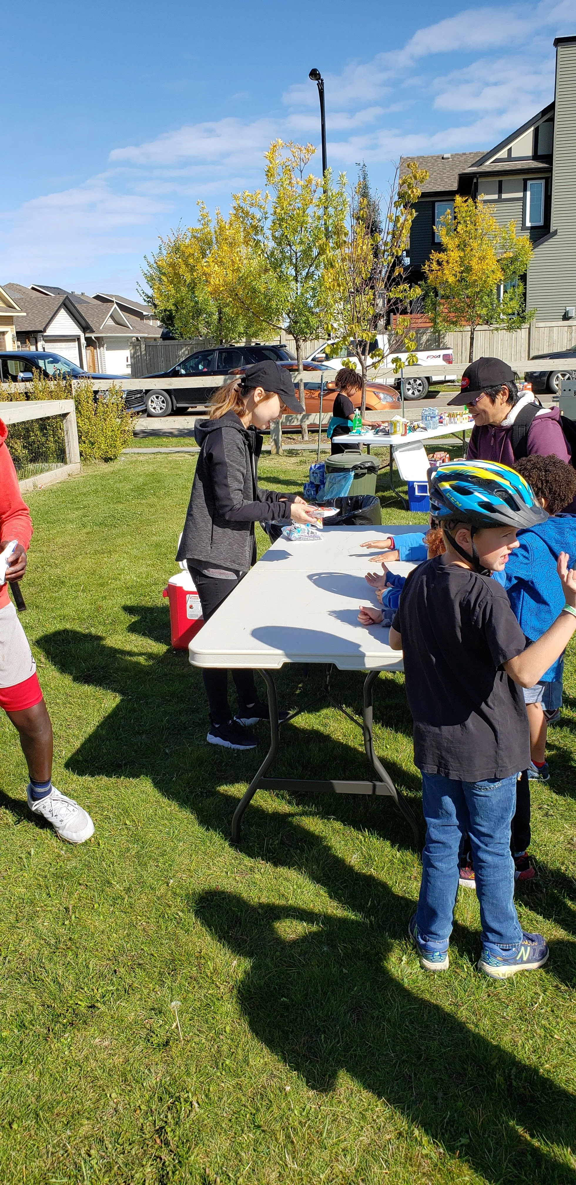 People standing at a table outside during a community event, with children and adults, near parked cars and residential houses.