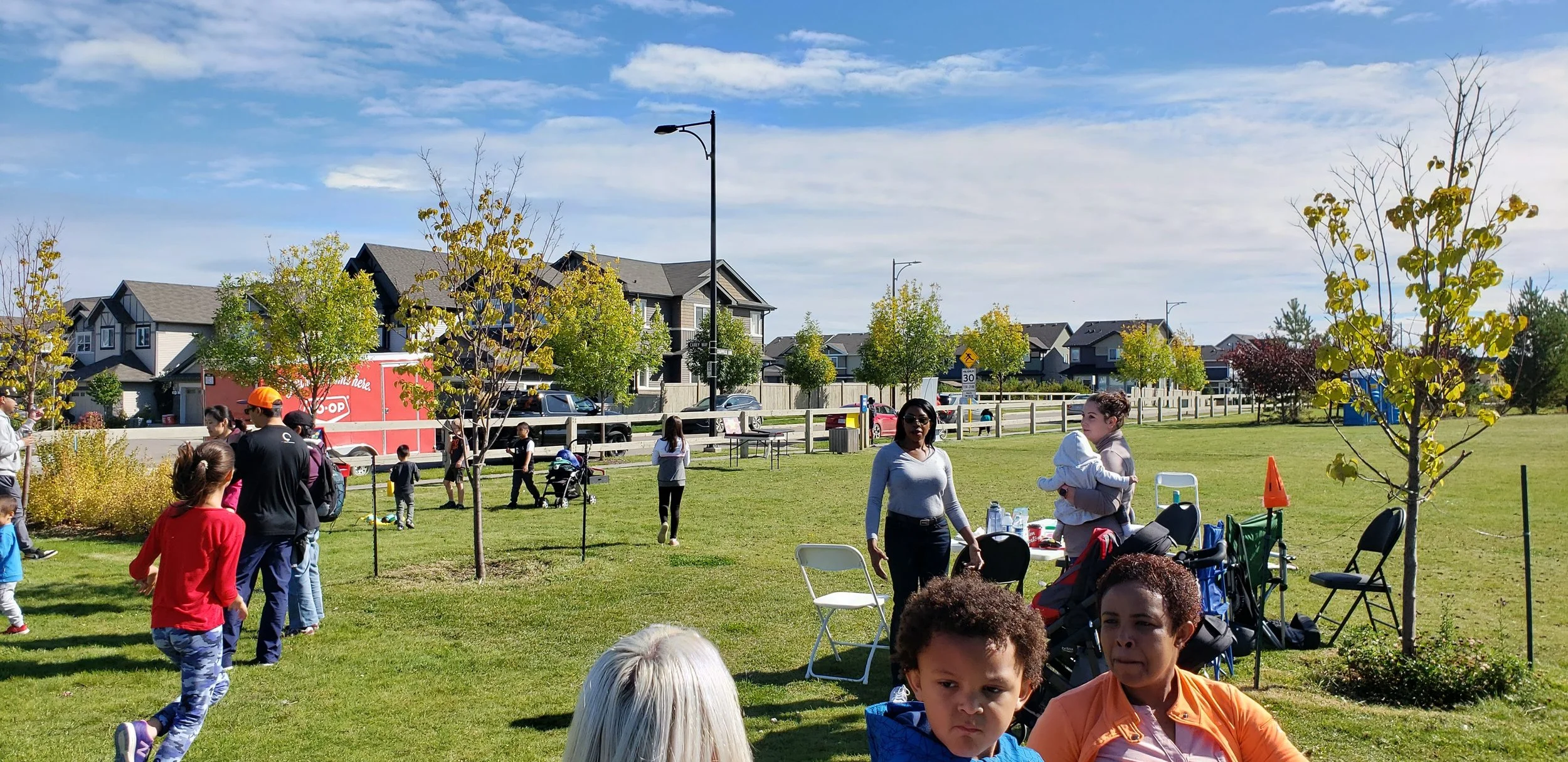 People gathered in a park on a sunny day, with children playing and adults socializing, surrounded by trees and residential houses in the background.