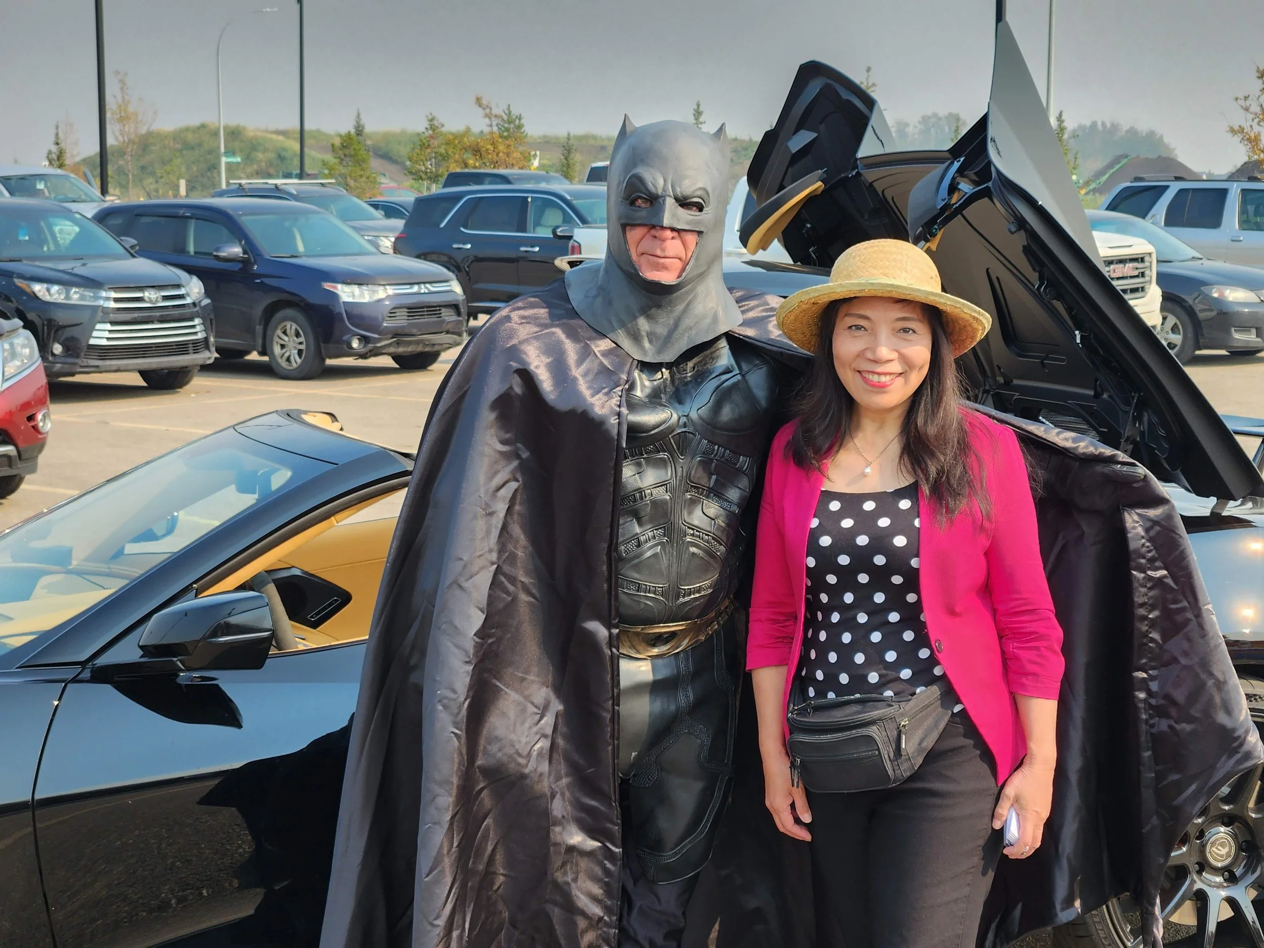 A person dressed as Batman and a woman in a pink blazer and straw hat standing beside a convertible car with the hood open in a parking lot.
