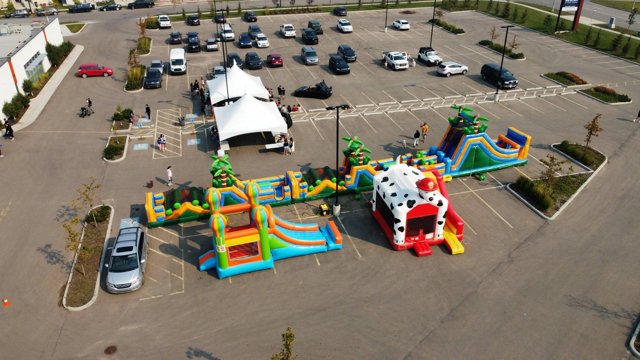 An outdoor event in a parking lot featuring inflatable bounce houses and slides decorated with a jungle theme and cow print. There are tents, children playing, and adults supervising, with cars parked around the area.