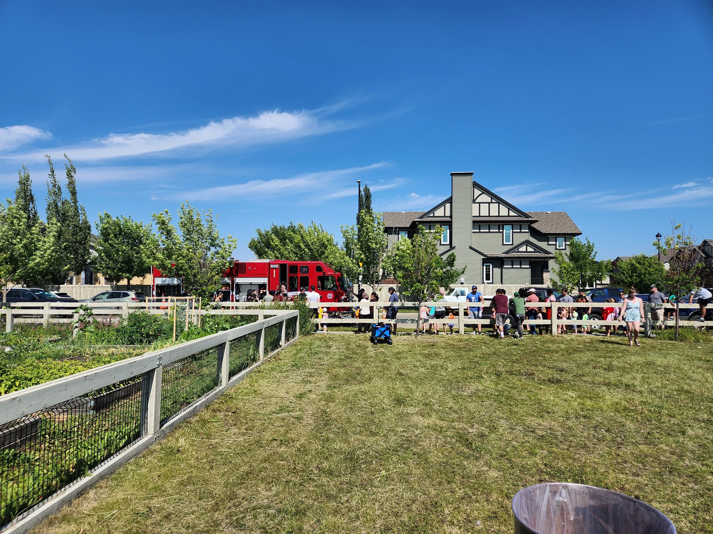 A group of people gathered outdoors behind a white fence, with a red fire truck parked in the background near a large, gray house and trees under a blue sky.