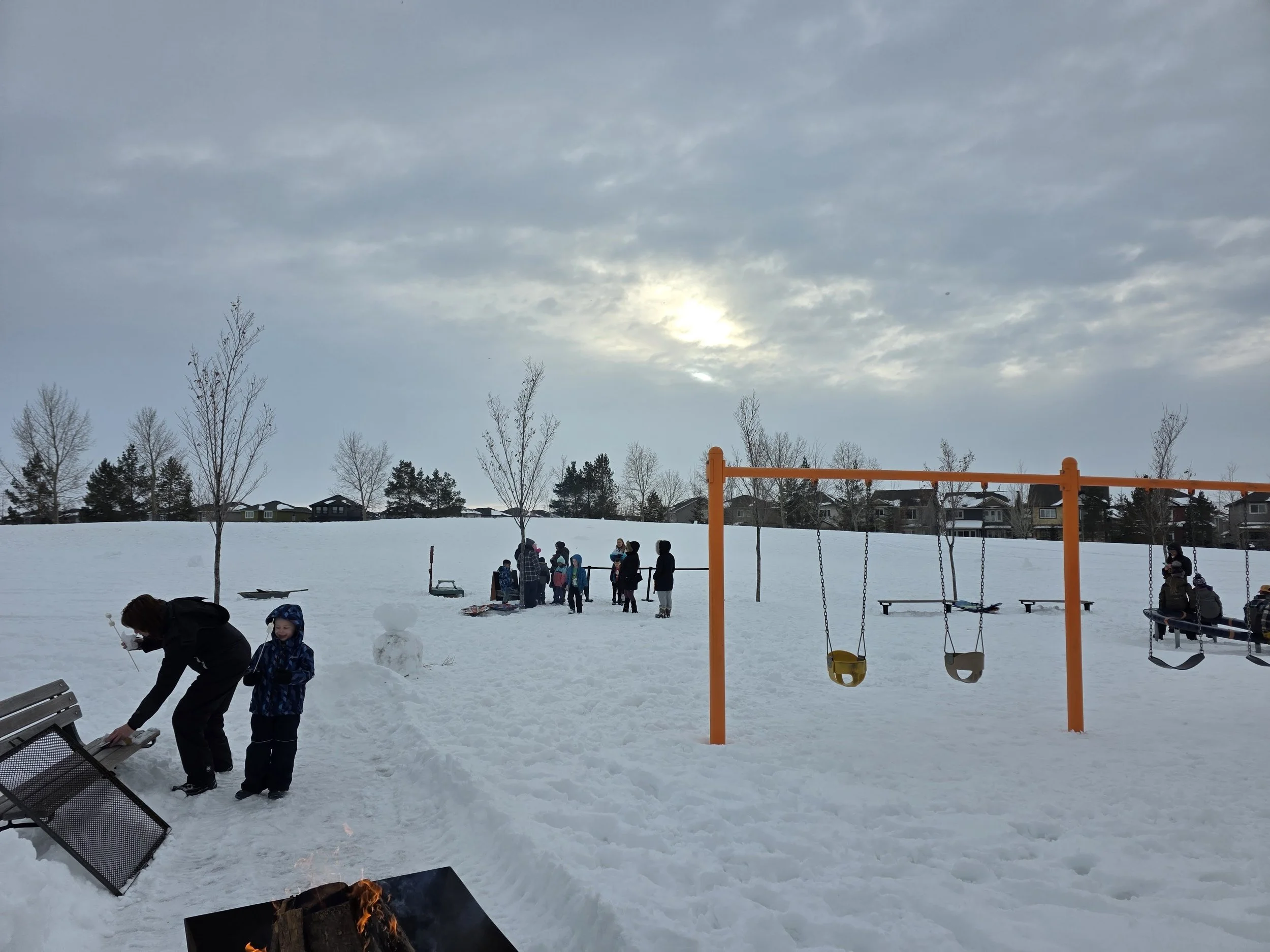 Children and adults gathering and playing in a snow-covered park with a swing set, benches, and a small snowman, under a cloudy sky during daytime.