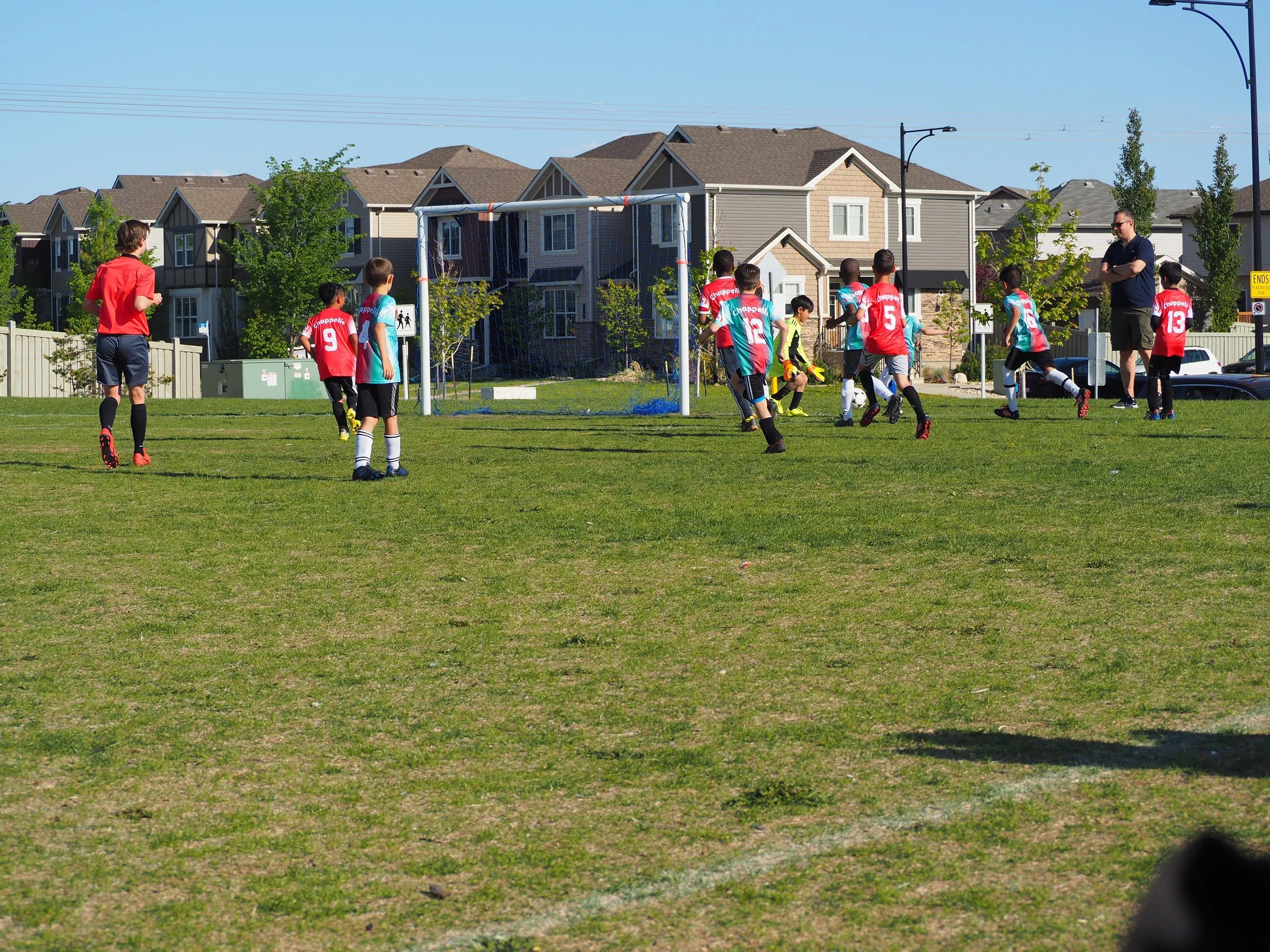 Children playing soccer on a field with two coaches, one in red and one in black, under a clear blue sky with residential houses in the background.