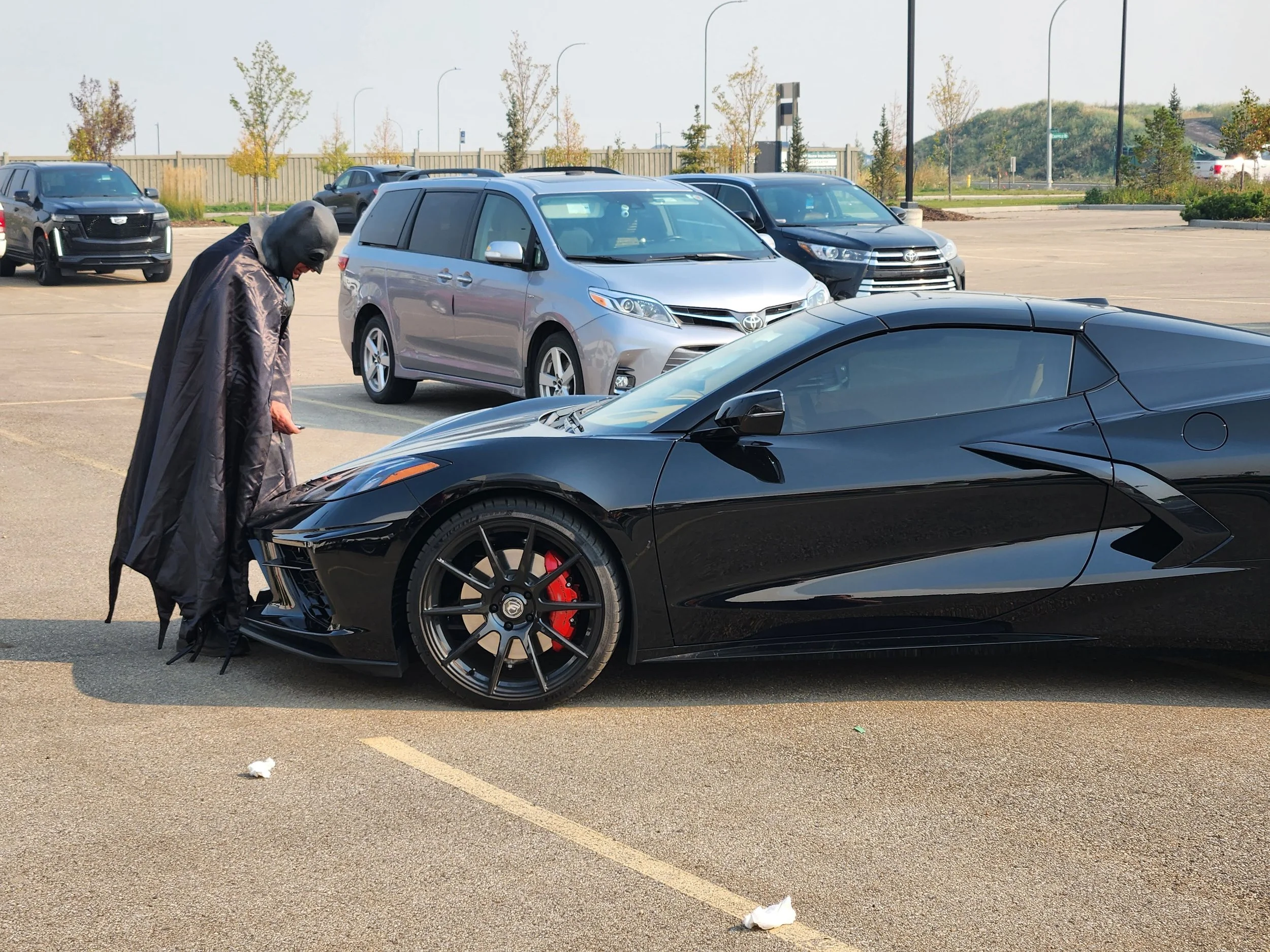 A person dressed as Batman looking at a black luxury sports car parked in an outdoor parking lot with other cars in the background.