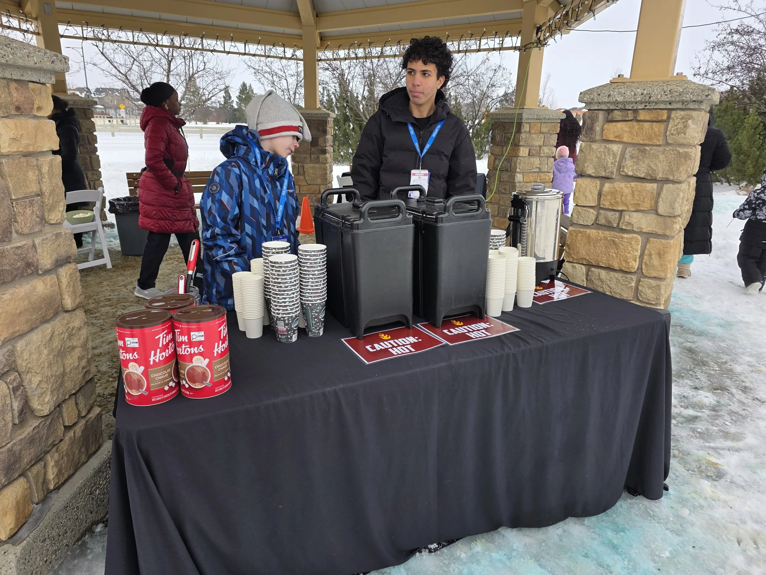 A food stand set up outdoors in a covered area with a black tablecloth. On the table are stacks of Tim Hortons hot chocolate cans, paper cups, and large beverage dispensers. Two people, a young boy in a blue jacket and a man in a black jacket, stand 