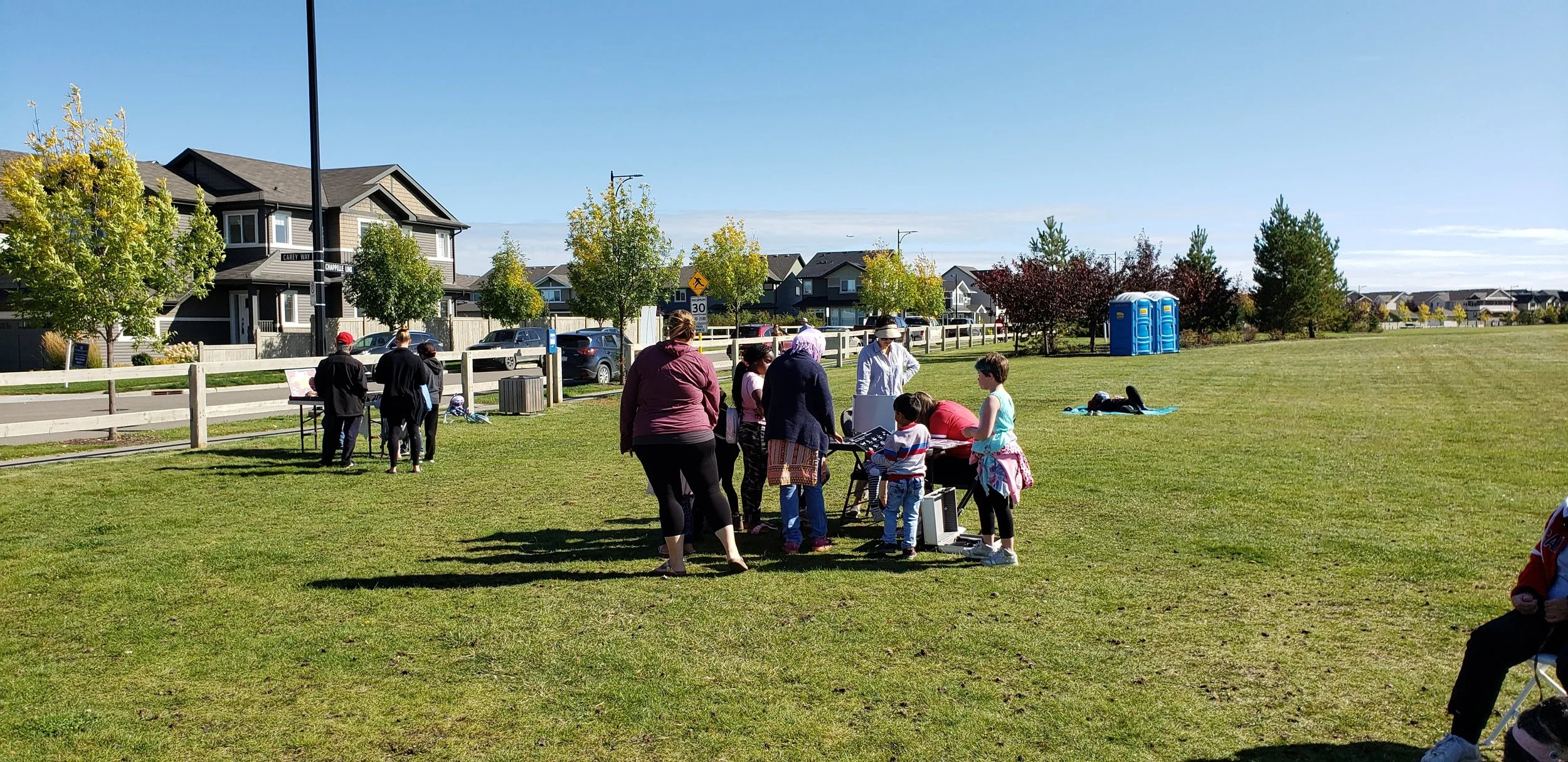 Group of people gathered in a park on a sunny day, some standing around a table, and others sitting or walking on the grass, with residential houses and trees in the background.