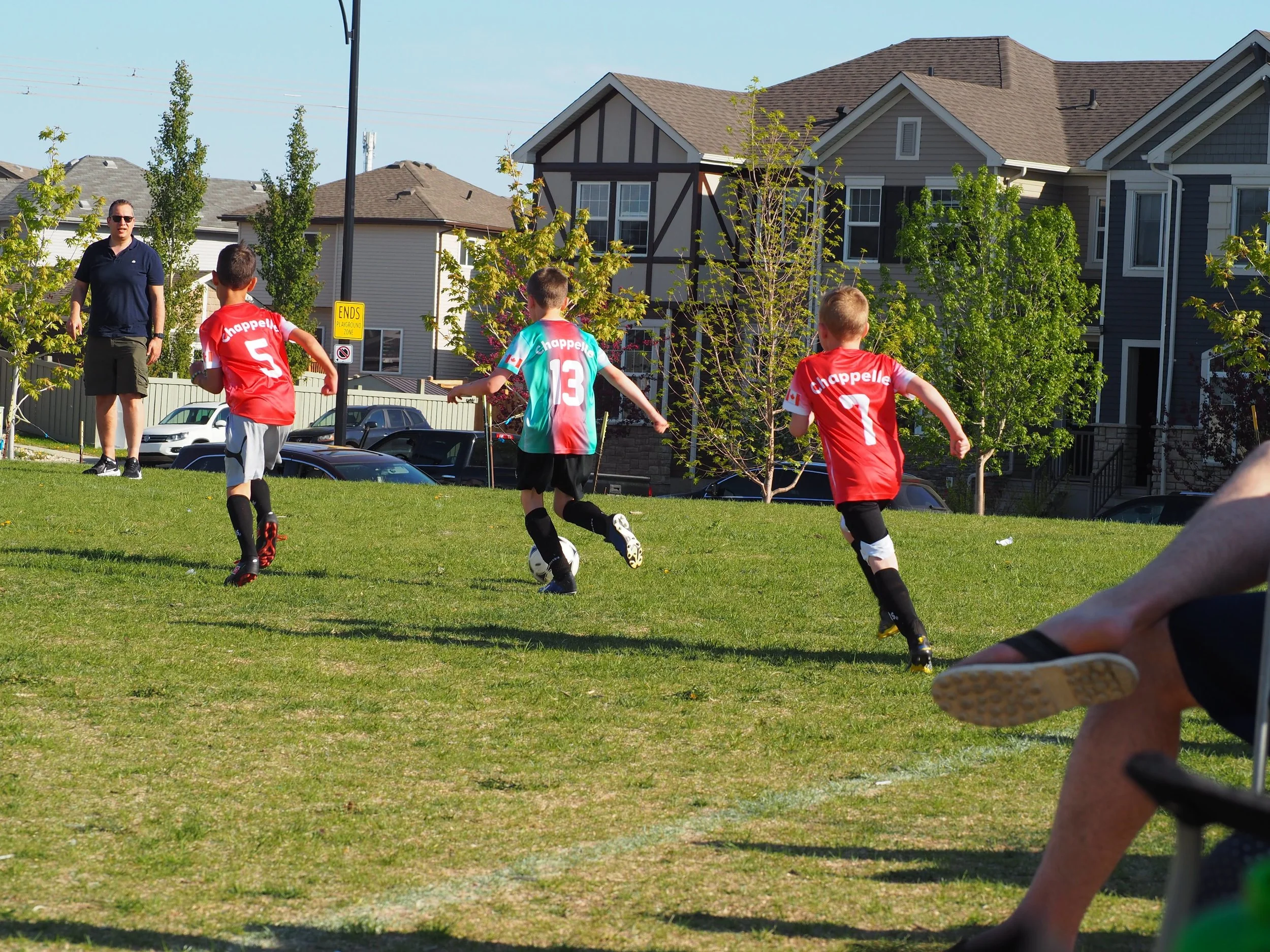 Children playing soccer on a grassy field with houses in the background.