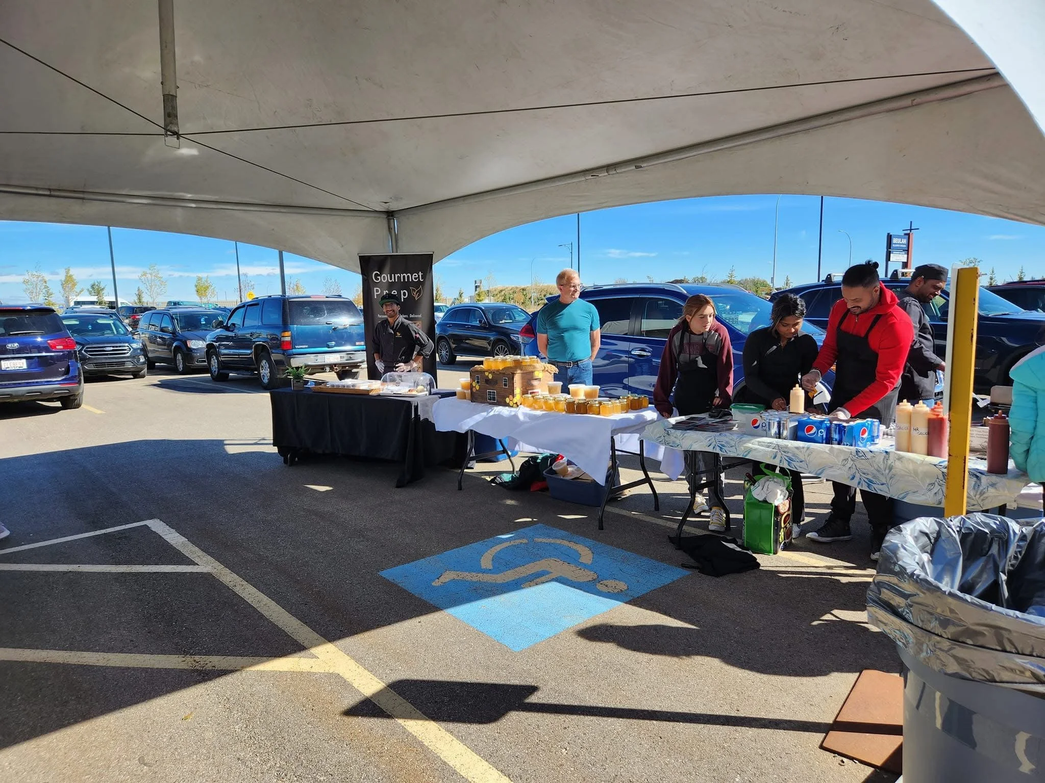 People at a food service table in a parking lot under a canopy, with parked cars behind them. The table has various food items and beverages, including mustard, ketchup, and soda, and a sign that reads "Gourmet Prep." A handicapped parking space is v