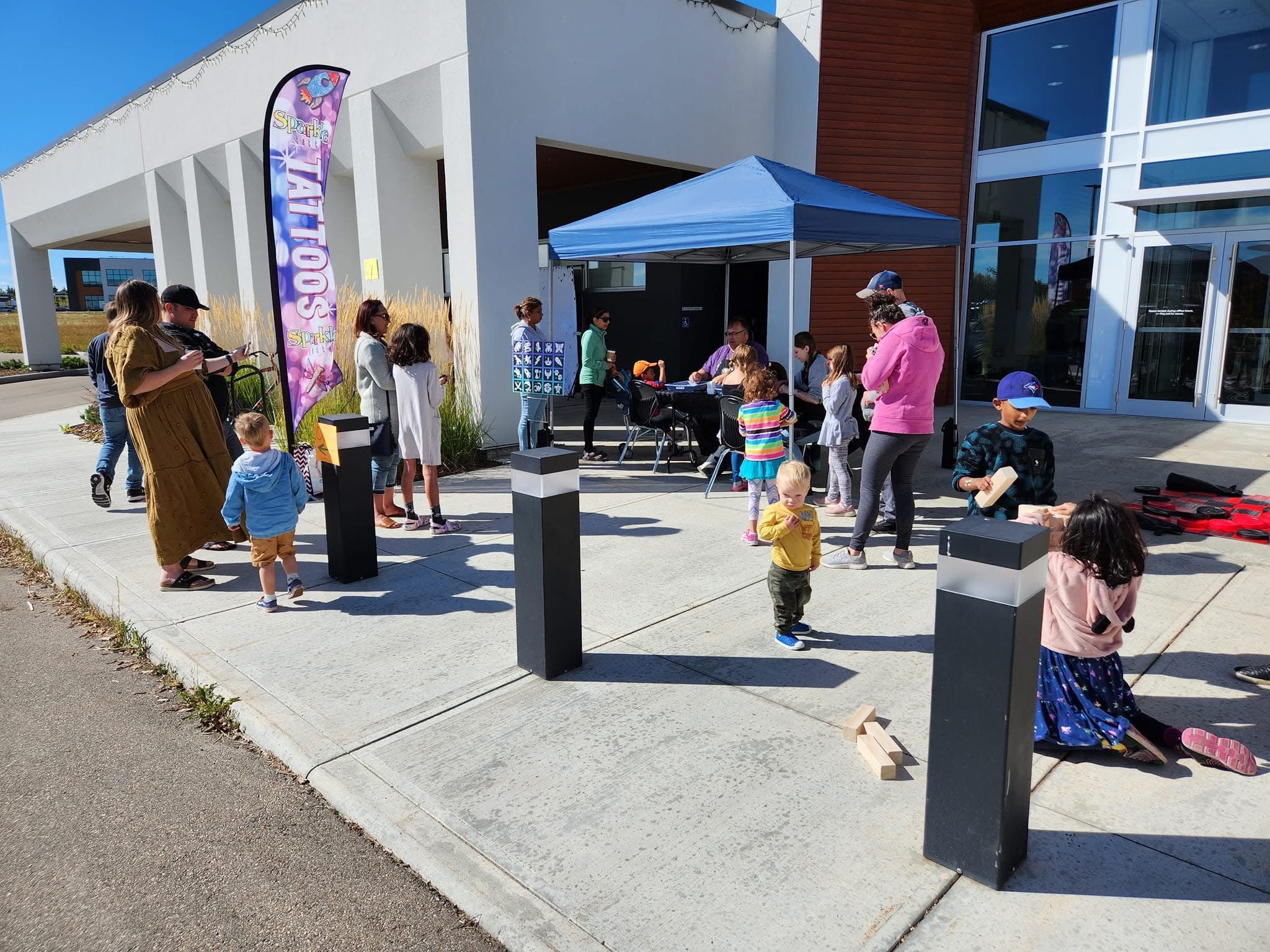 Children and adults participating in an outdoor event at a modern building with a Sparkle Tattoos banner, some are playing games, standing in line, or sitting at tables under a blue canopy.