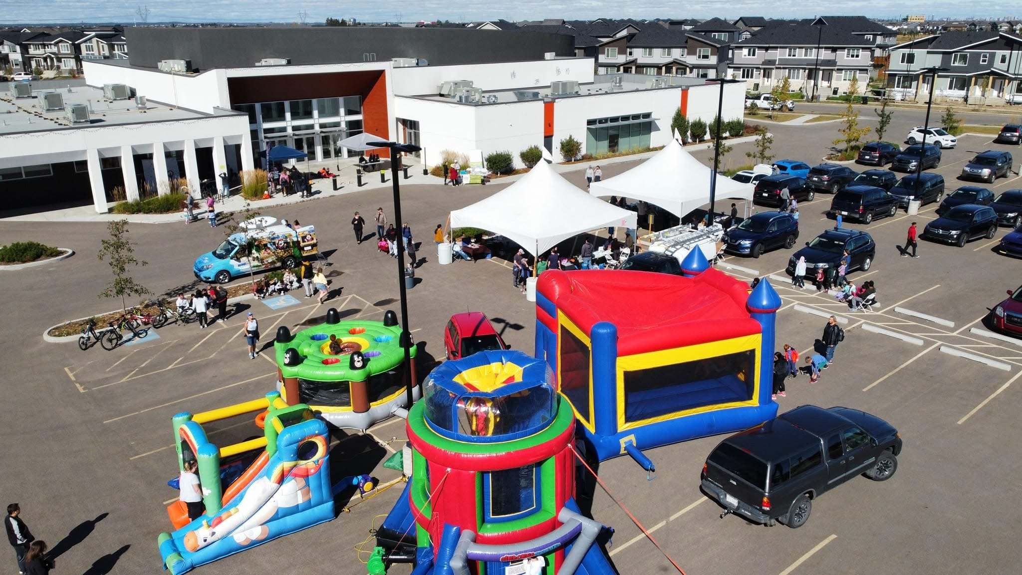 Parking lot outside a community center with inflatable bounce houses, a small amusement ride, tents with vendors, and people walking and gathered around.