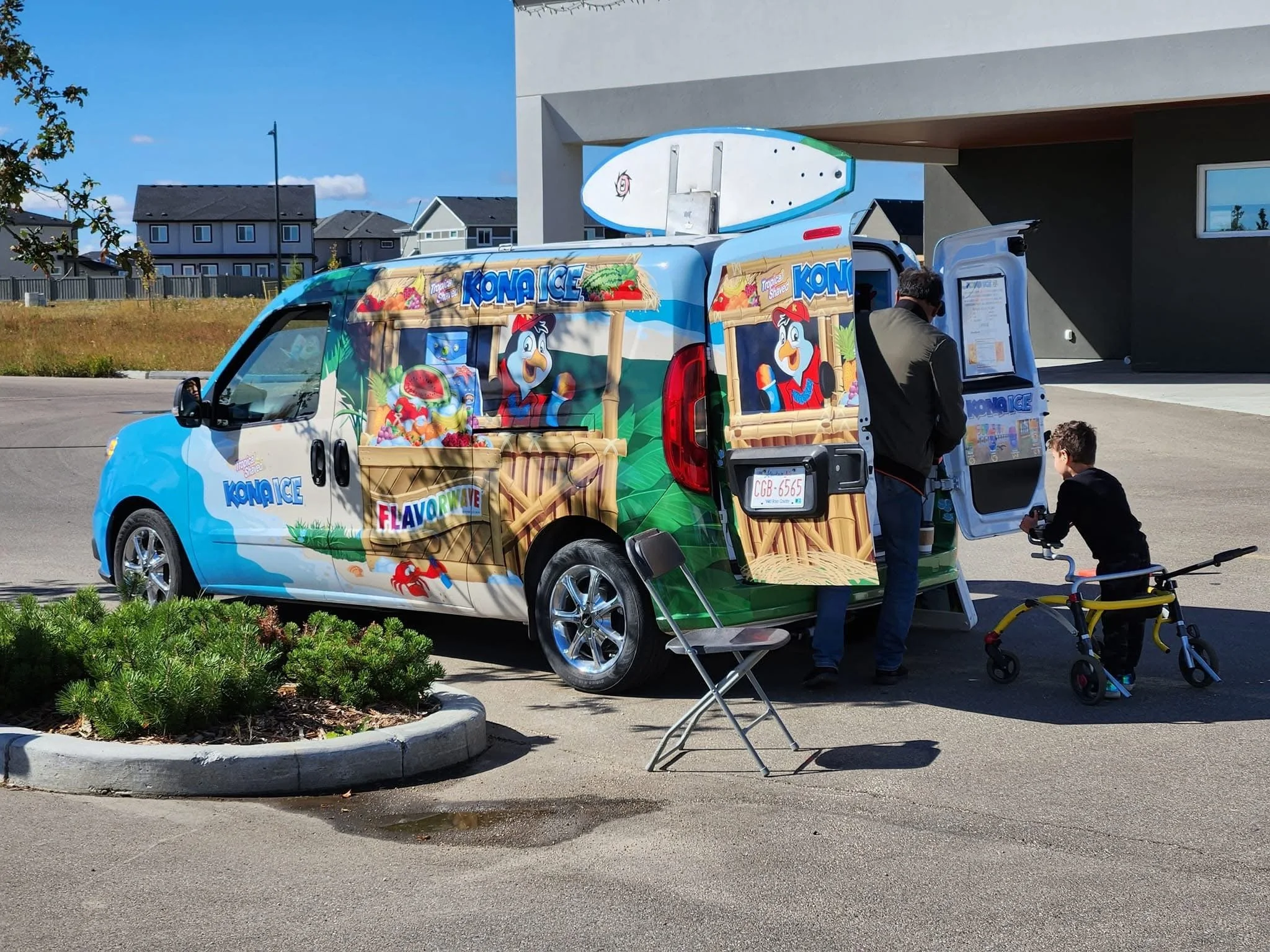 A colorful Kona Ice snow cone truck parked outside. Two boys are ordering at the window, with one boy standing on a yellow bicycle nearby. The truck has images of a cartoon penguin and tropical flavors on its sides.