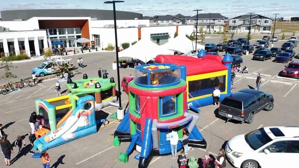 Children playing on colorful inflatable bounce houses in a parking lot outside a shopping mall, with cars parked around and people walking.