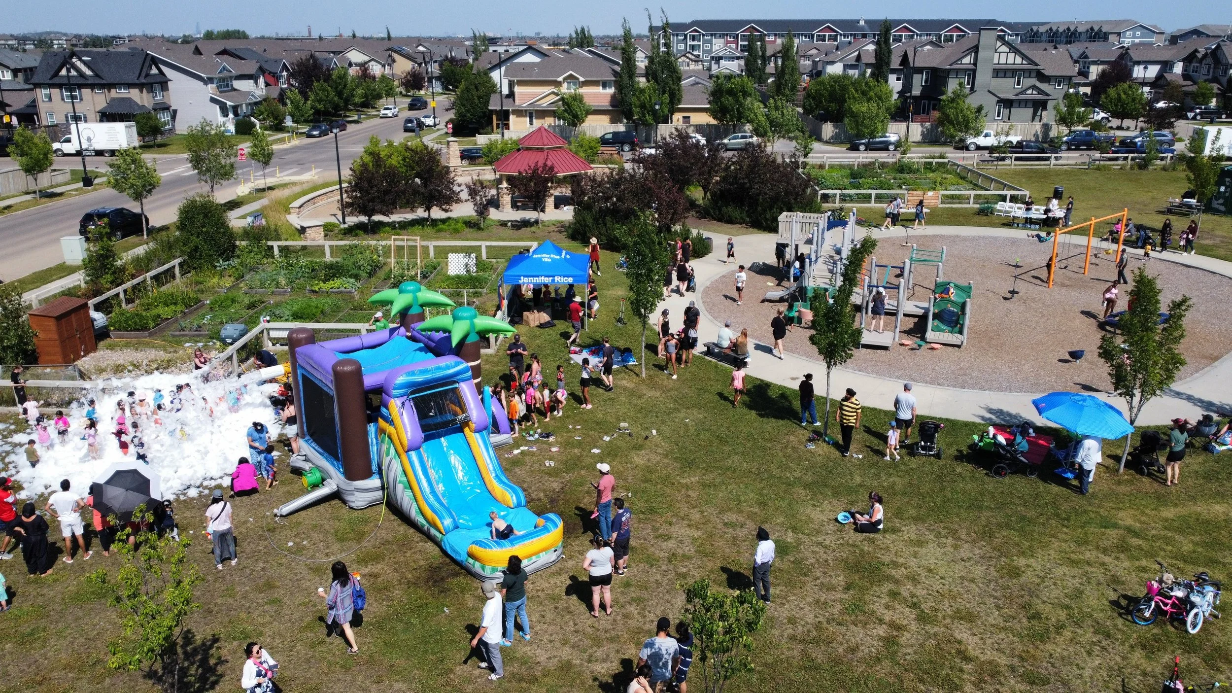 A community park with children playing on an inflatable water slide and water foam area, adults watching under umbrellas, and a playground in the background. Houses and a street are visible beyond the park.