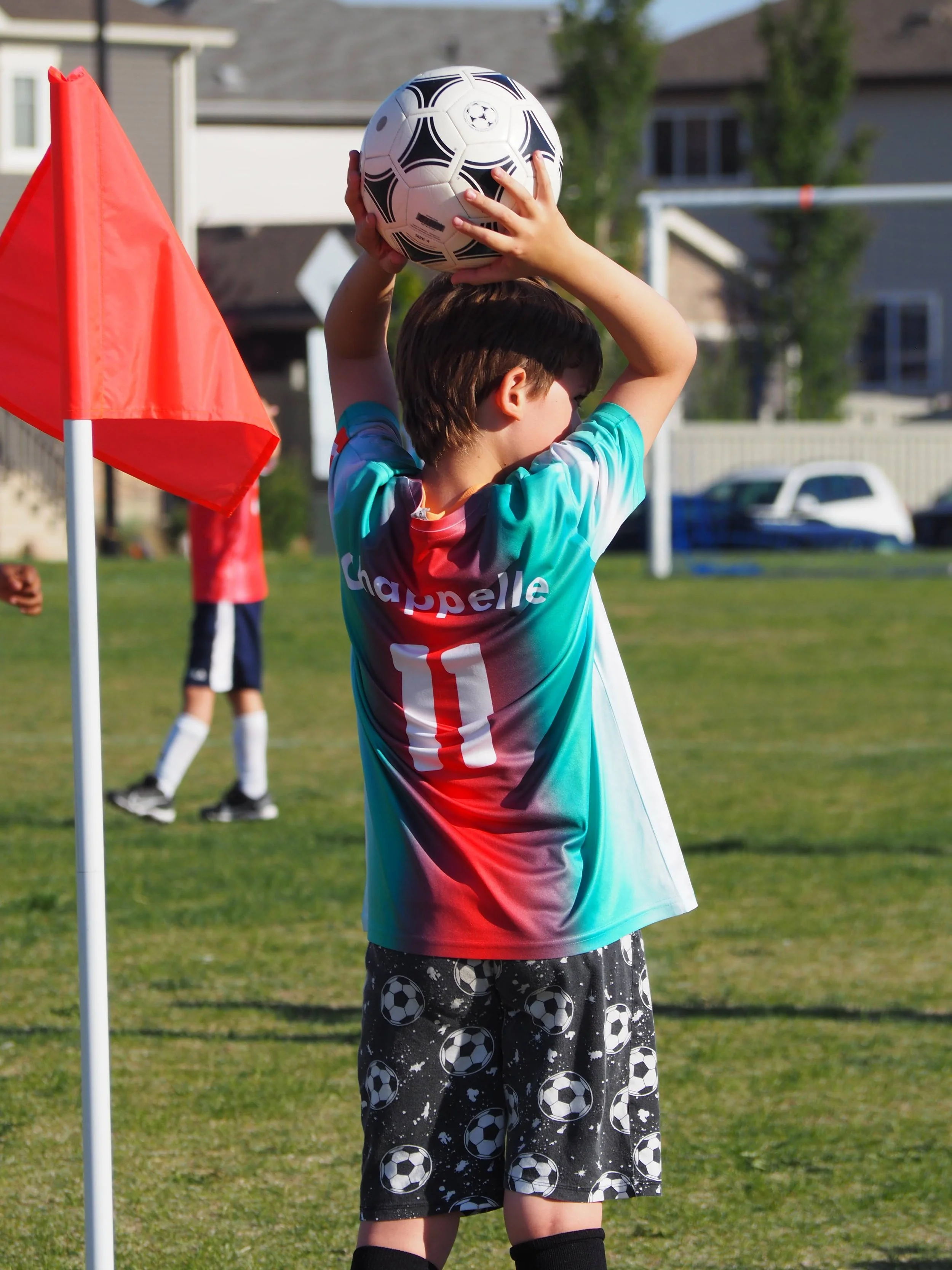 Child playing soccer, holding a soccer ball above their head, on a grassy field with houses in the background, near a red corner flag.