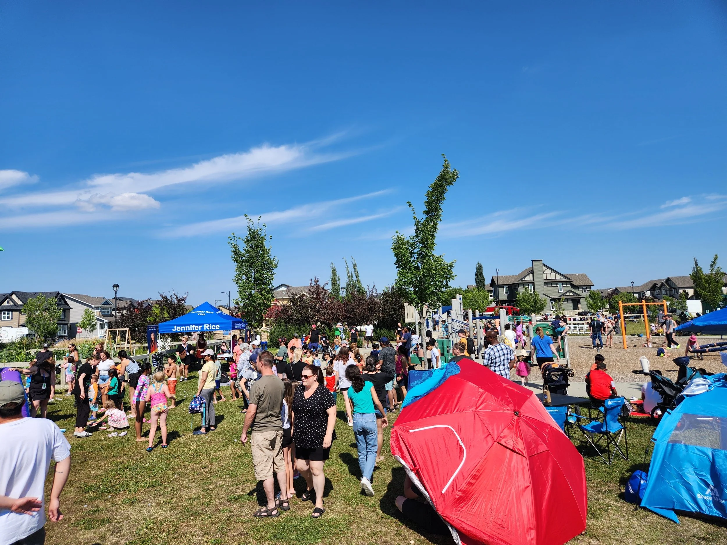 A crowded outdoor park scene with many people, tents, and playground equipment under a blue sky with clouds.