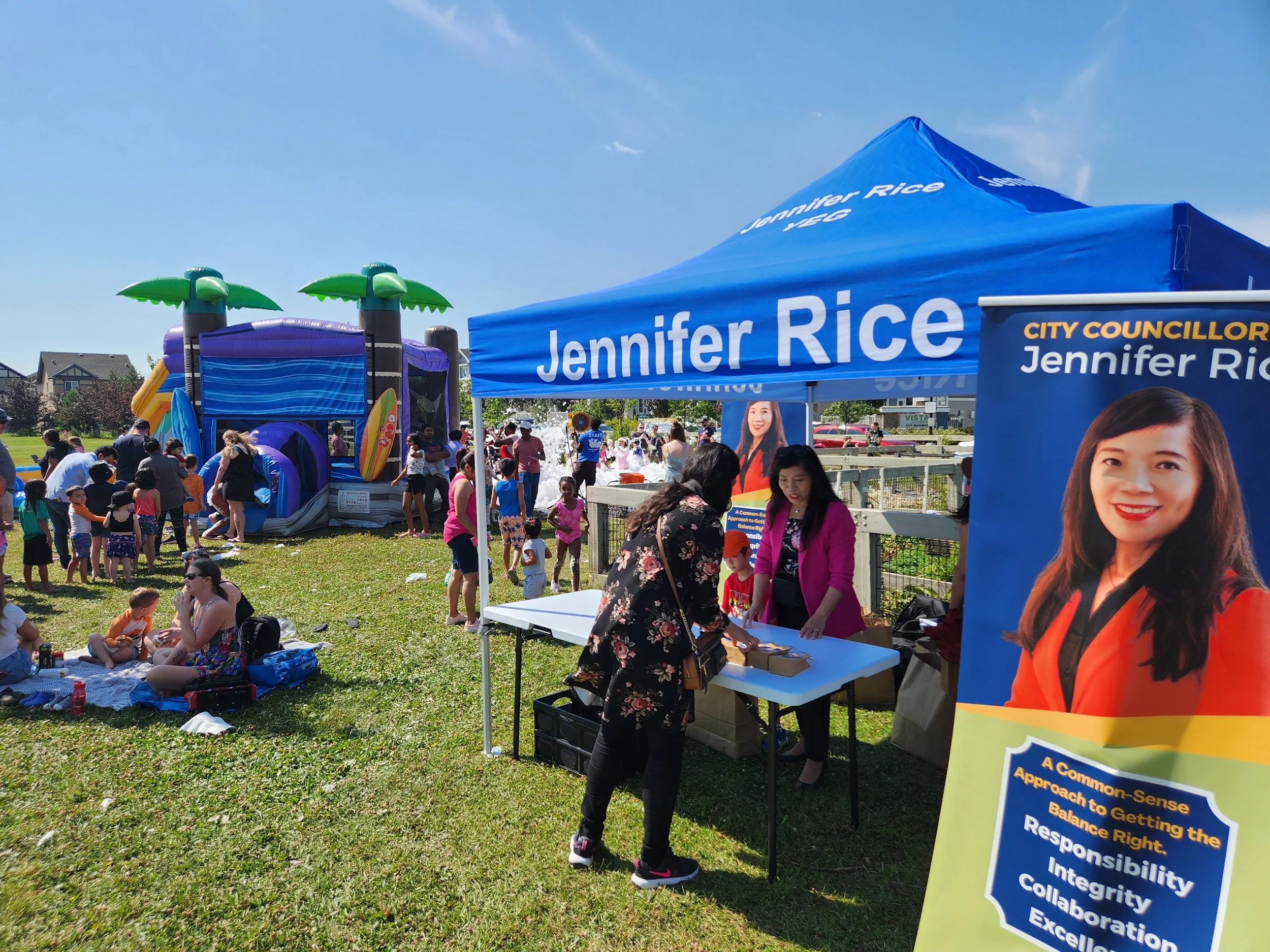 A community outdoor event featuring children playing on a large inflatable water slide with palm tree decorations, people sitting on the grass, and two women at a table under a blue Jennifer Rice campaign tent.