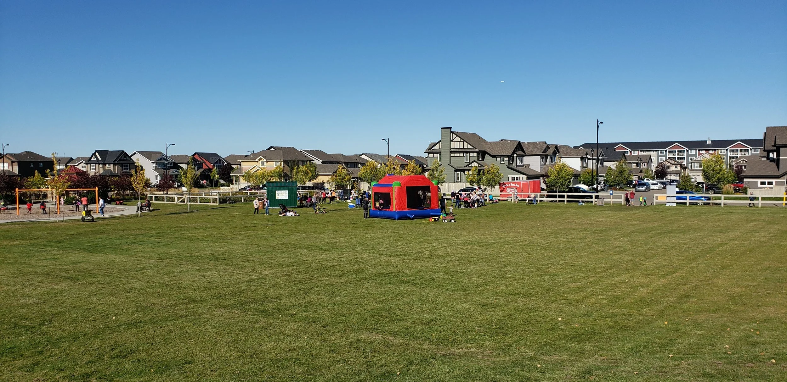 A community park on a clear day with people gathered around a colorful inflatable bounce house, children playing on a swing set, and houses in the background.
