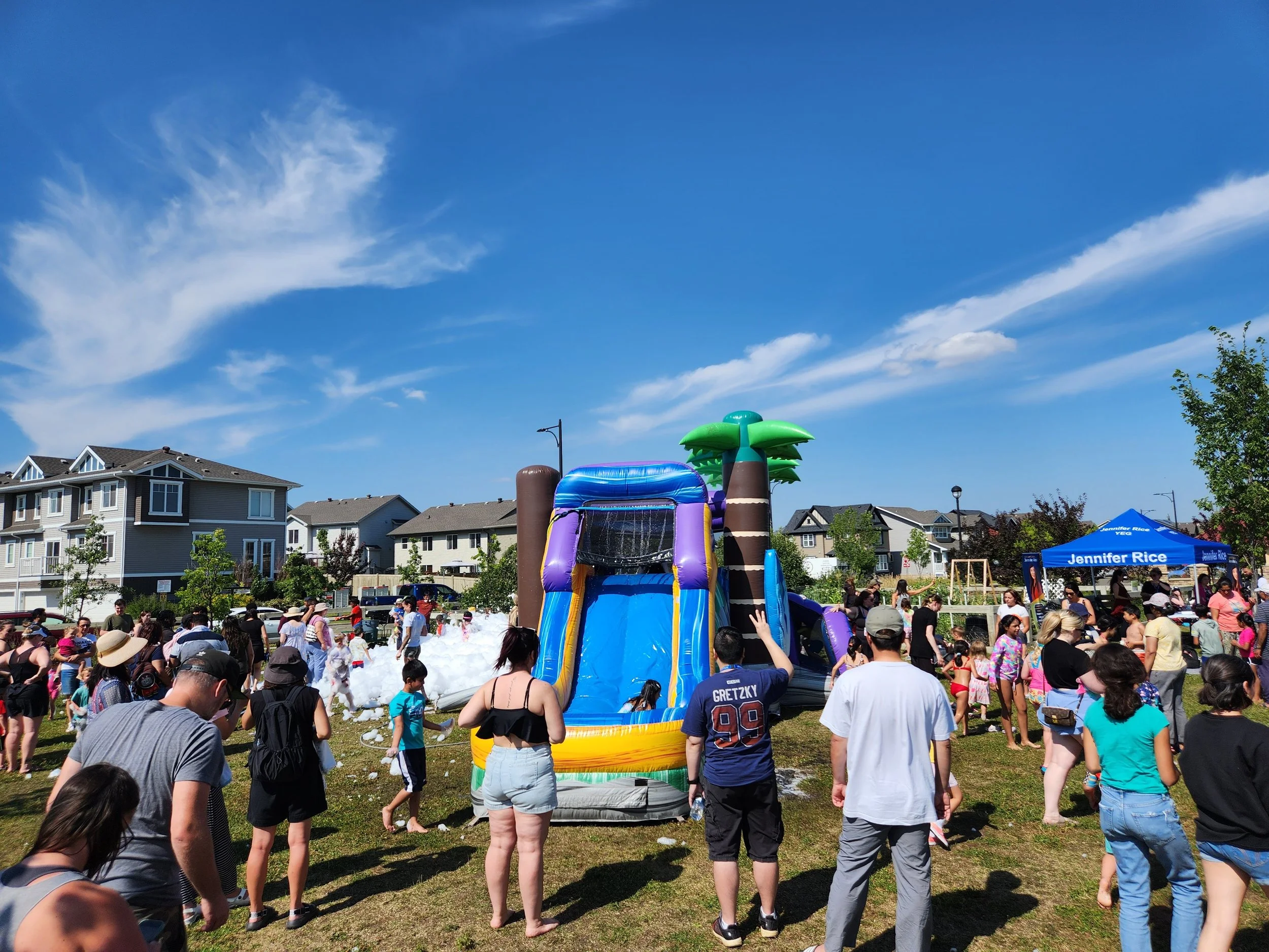 People gathered around an inflatable water slide with a palm tree design in an outdoor park on a sunny day