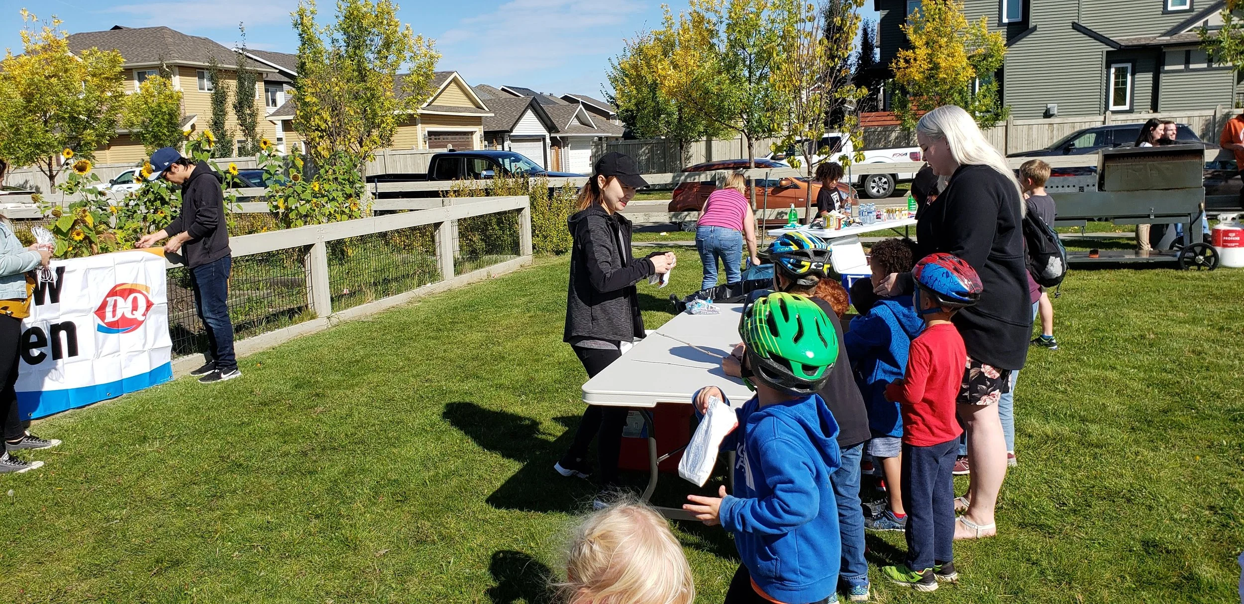 Children and adults gather outdoors at a community event near a table with ice cream, wearing helmets, on a sunny day with houses, parked cars, and trees in the background.