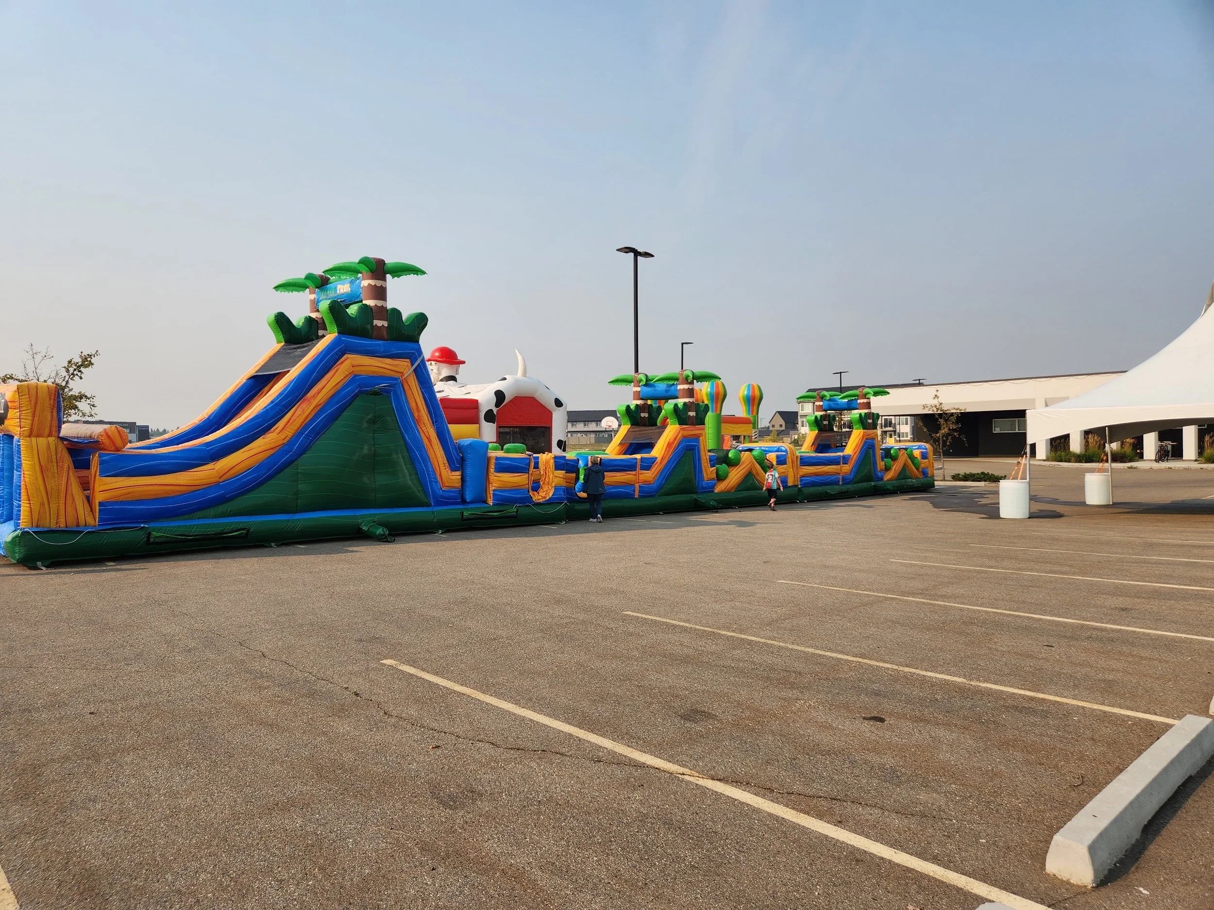 Colorful inflatable kiddie park with slides, palm trees, and a cow-themed bounce house in an empty parking lot.