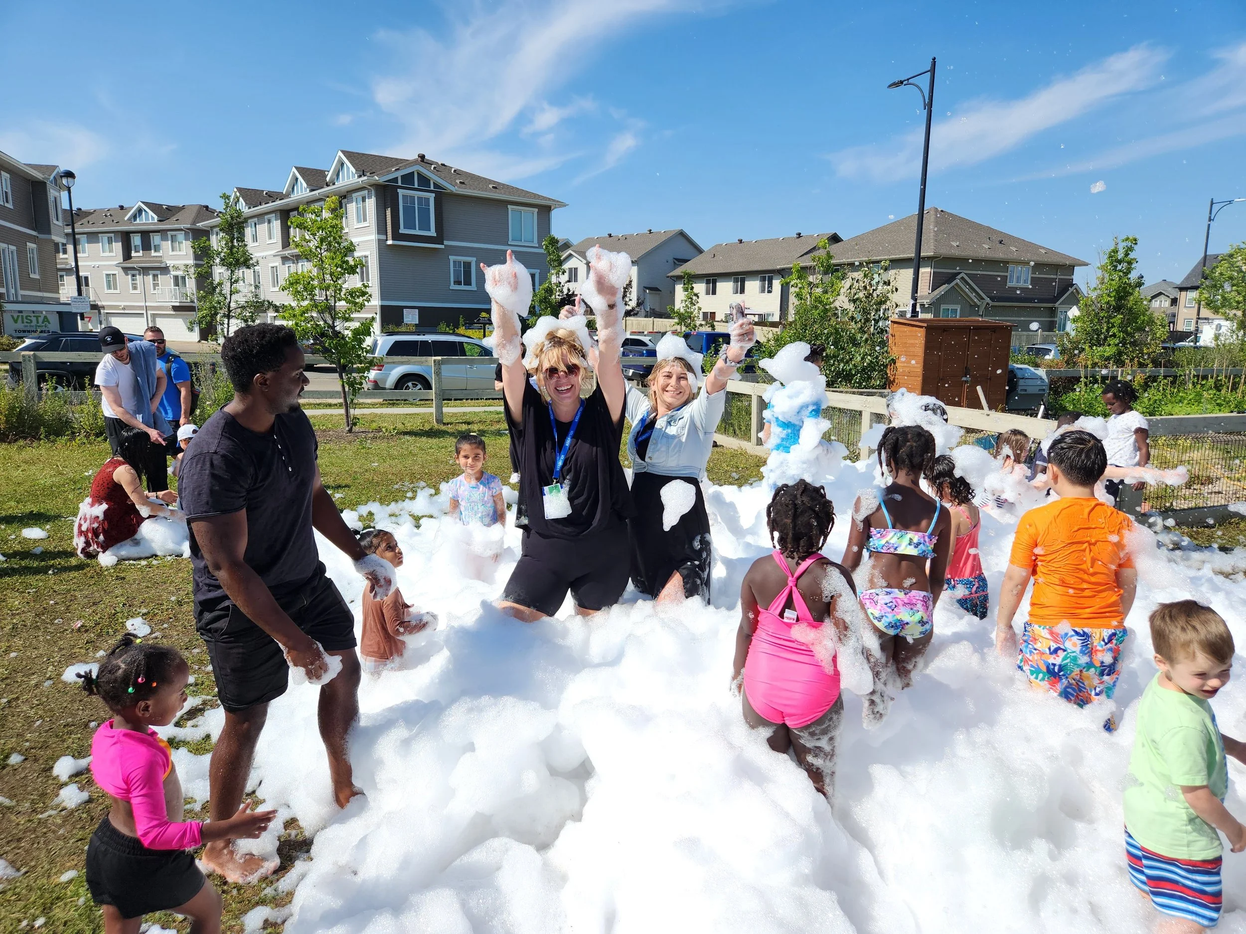 Children and adults playing in foam on a grassy area under a bright blue sky, surrounded by trees and modern houses.