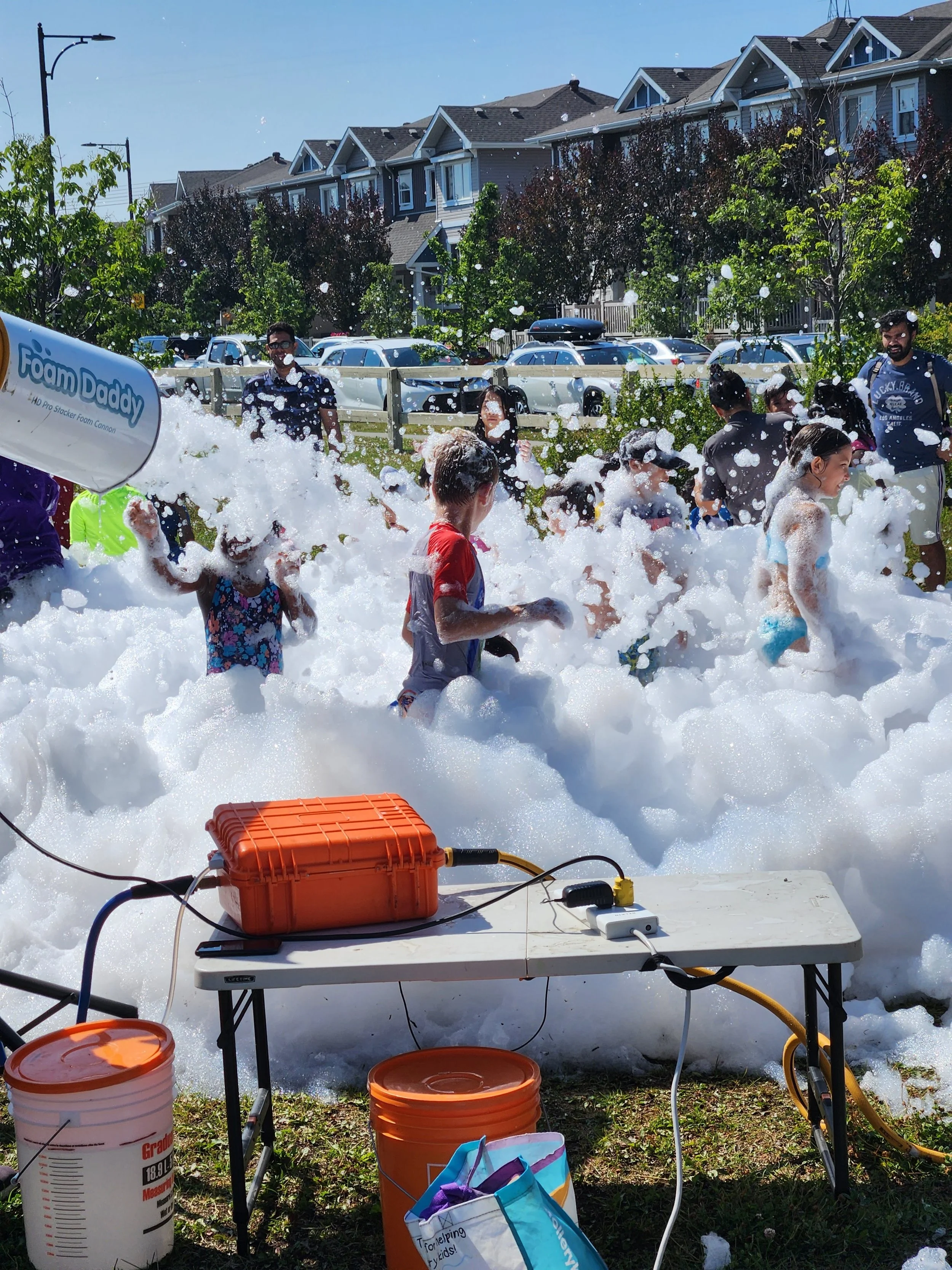 Children and adults playing in foam at an outdoor event on a sunny day, with foam machine in use and foam reaching the ground, surrounded by houses and parked cars.