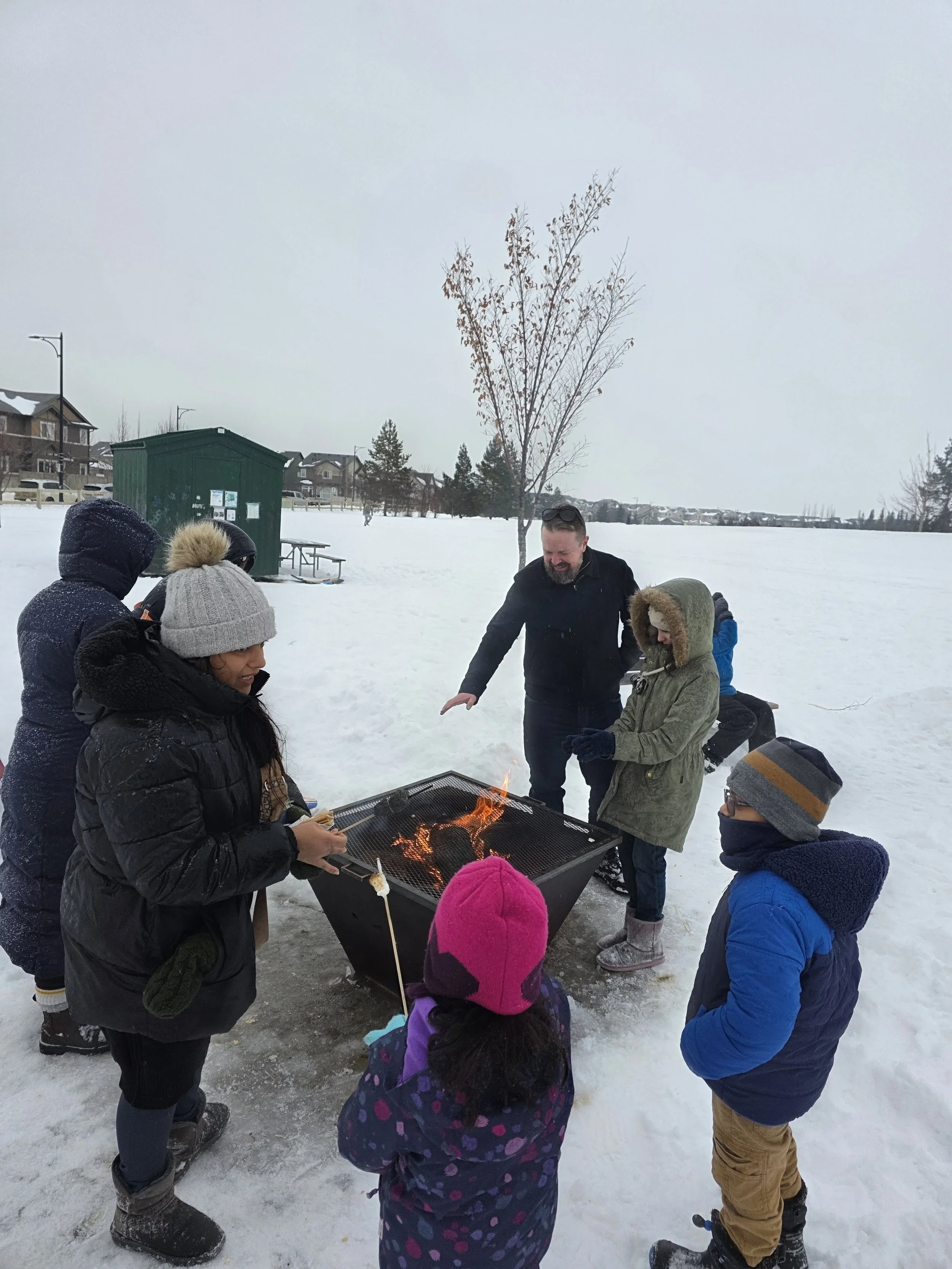 Group of children and adults gathered around an outdoor grill in snowy park, roasting marshmallows over an open flame.