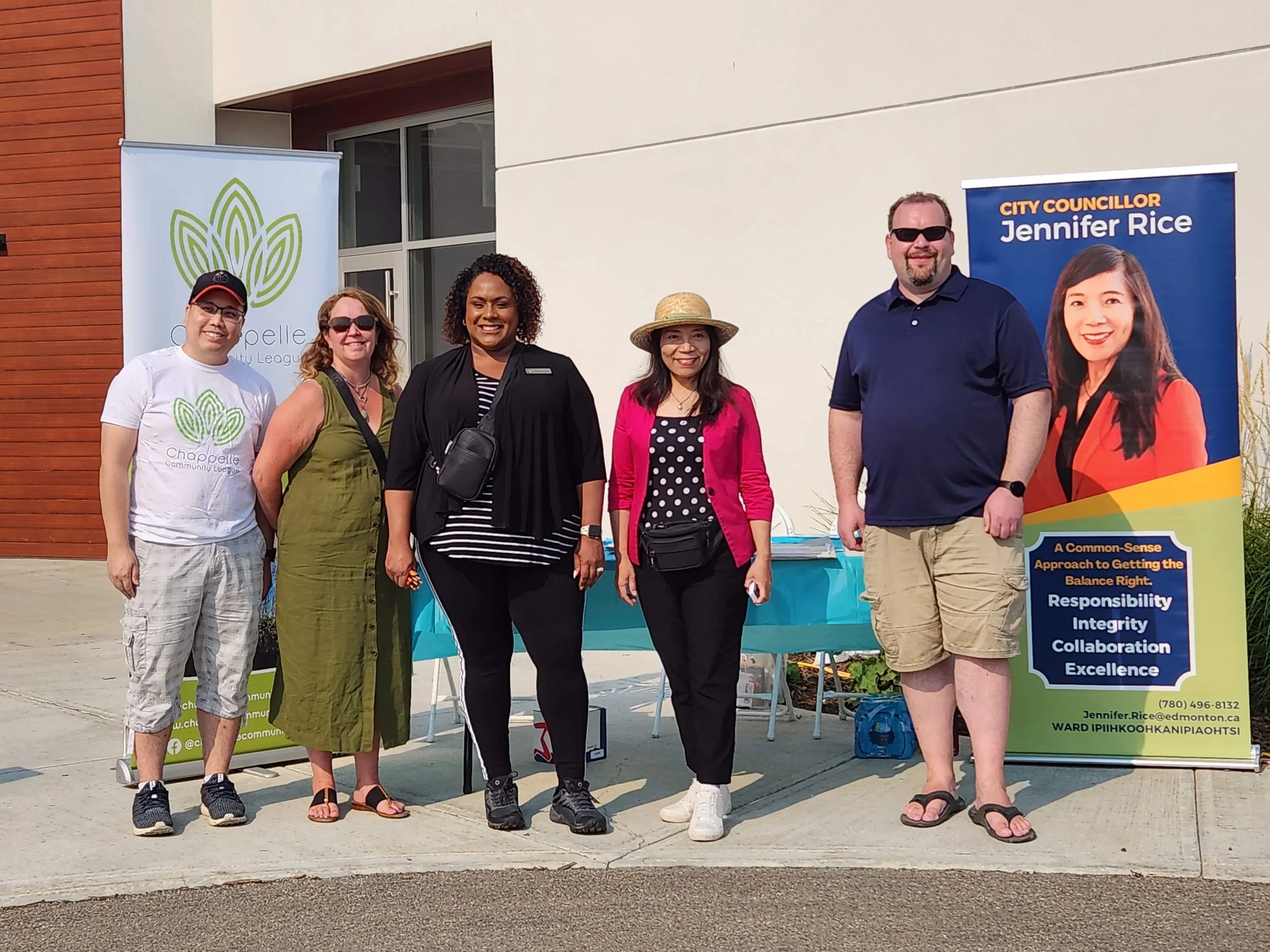 Group of five people standing outdoors in front of two banners, one with a green logo and the other with a photo of a woman and the name Jennifer Rice, during a community event.