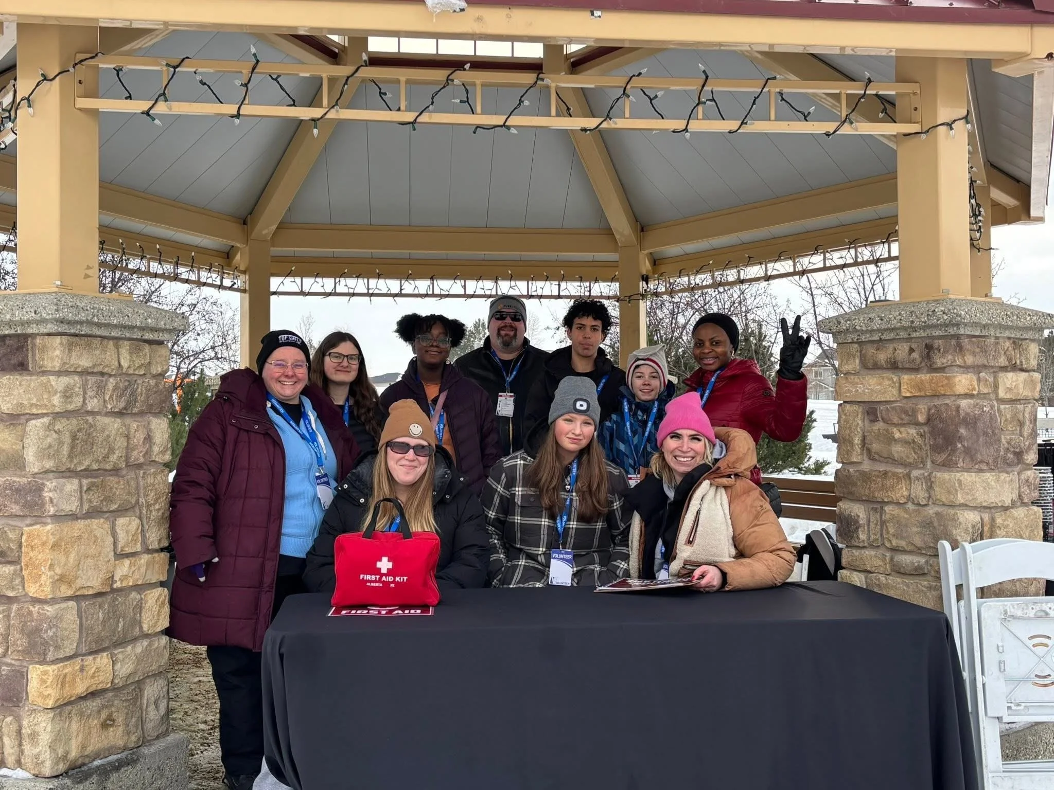 Group of people, both children and adults, gathered under a gazebo with stone supports and string lights. They are smiling, some making peace signs, and are wearing winter clothing including hats and jackets. There is a table with a first aid kit and