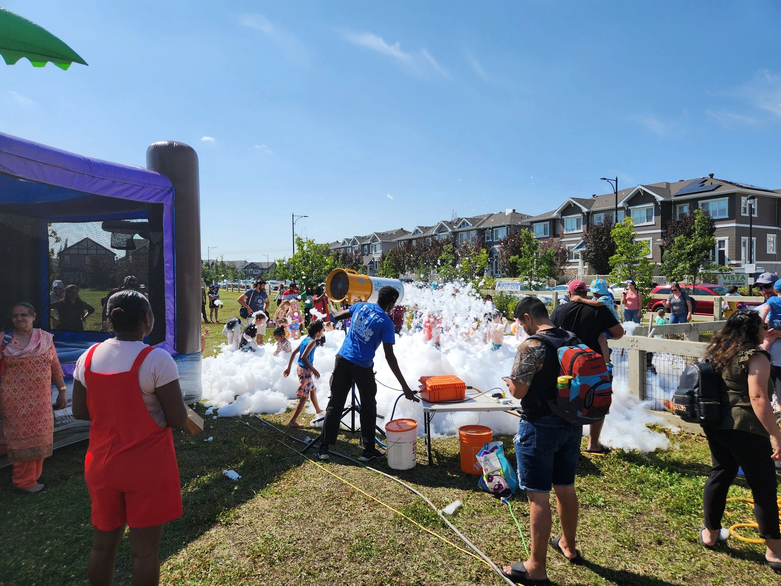 People having fun at a foam party outdoors on a sunny day, with children and adults playing in foam near a bounce house and a foam machine, with residential houses in the background.