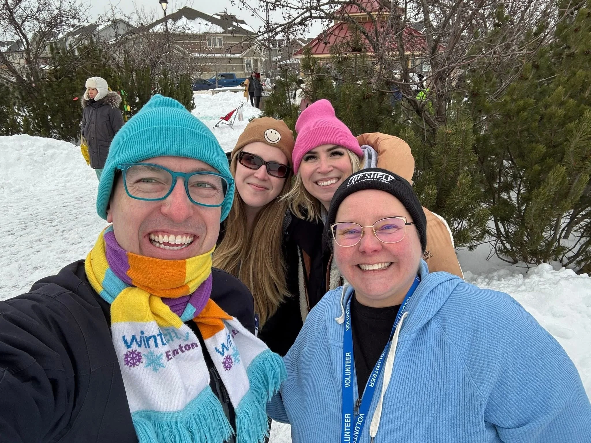 Group of four smiling friends taking a selfie outdoors in snowy winter setting, wearing colorful winter clothing and accessories, with other people and snow-covered trees and houses in the background.