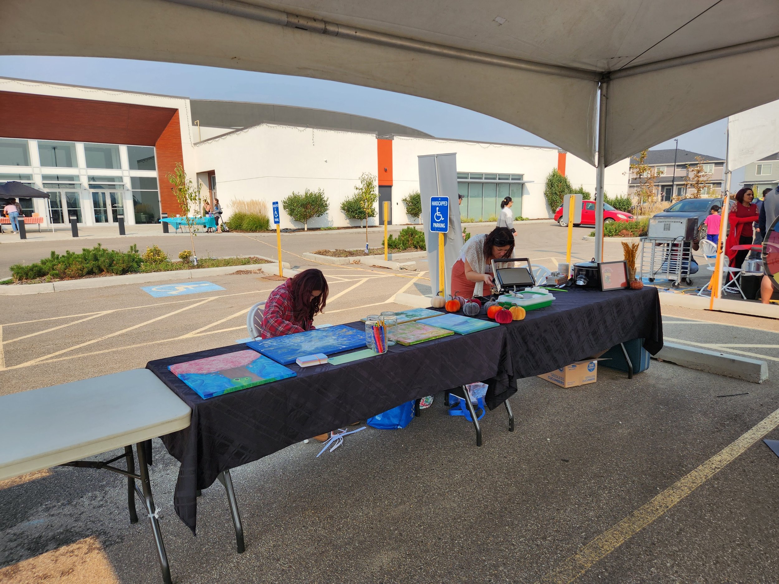 Outdoor craft fair with two women working at a table under a tent, displaying paintings, craft supplies, and fabric pumpkins.