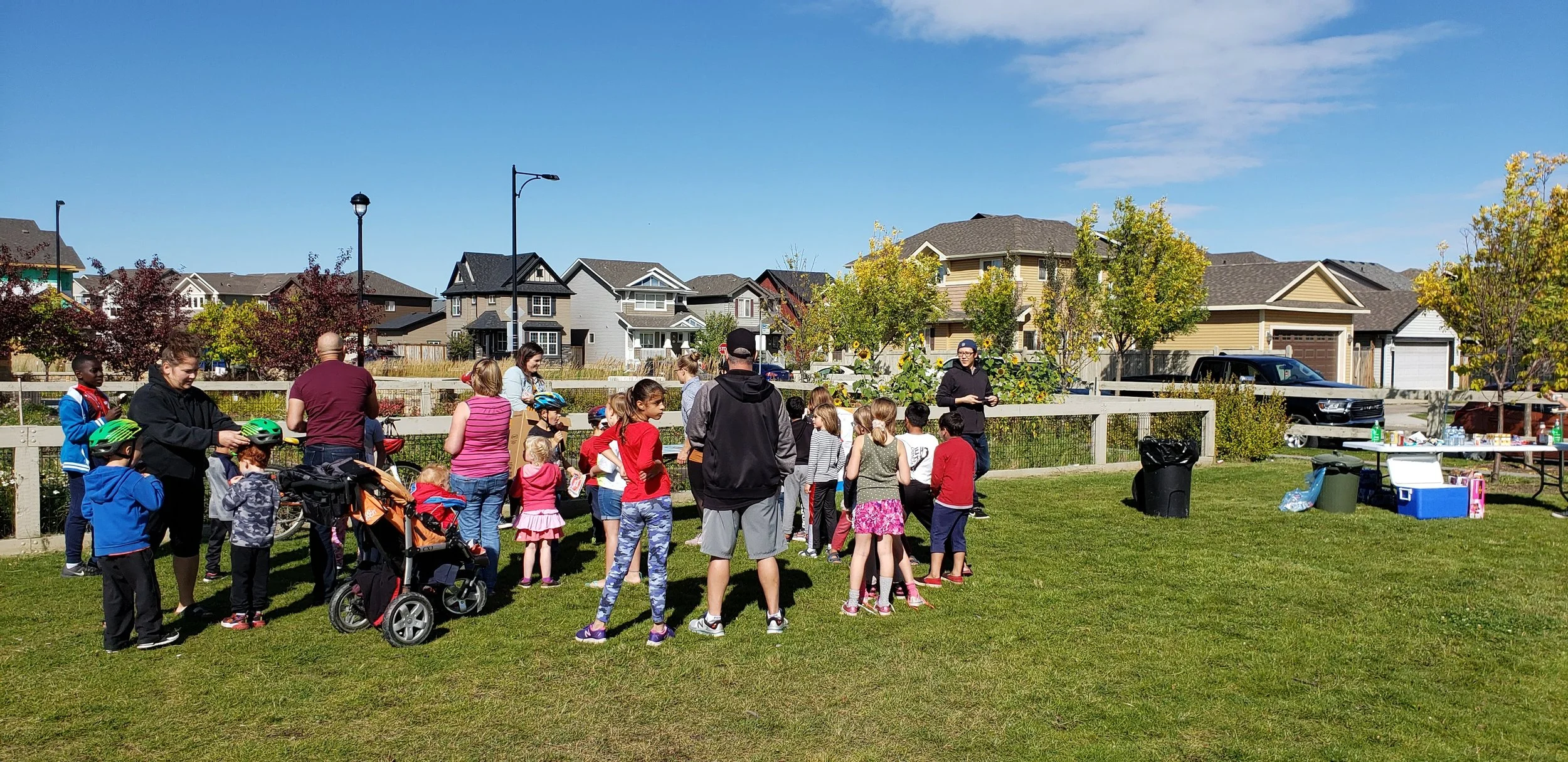 A group of children and adults gathered outdoors on a grassy field for an event in a residential neighborhood, with tables, coolers, and vehicles in the background.