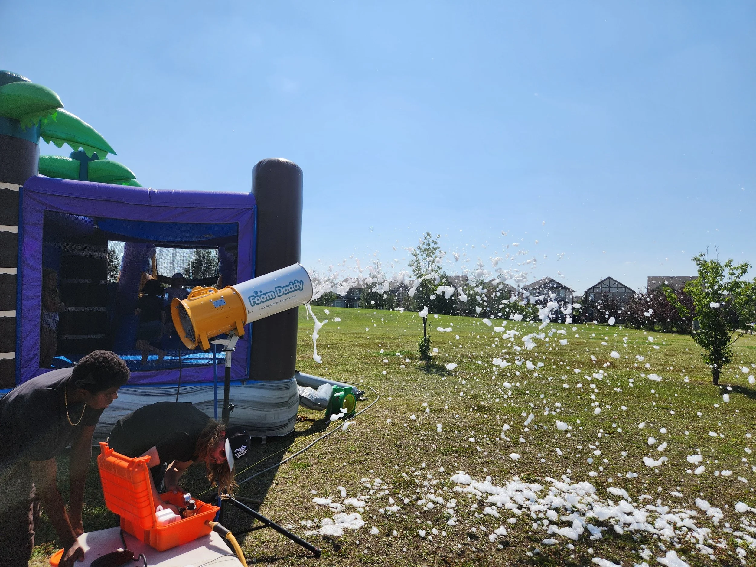 Kids playing outside with a foam machine shooting foam bubbles into the air near an inflatable bounce house on a sunny day.