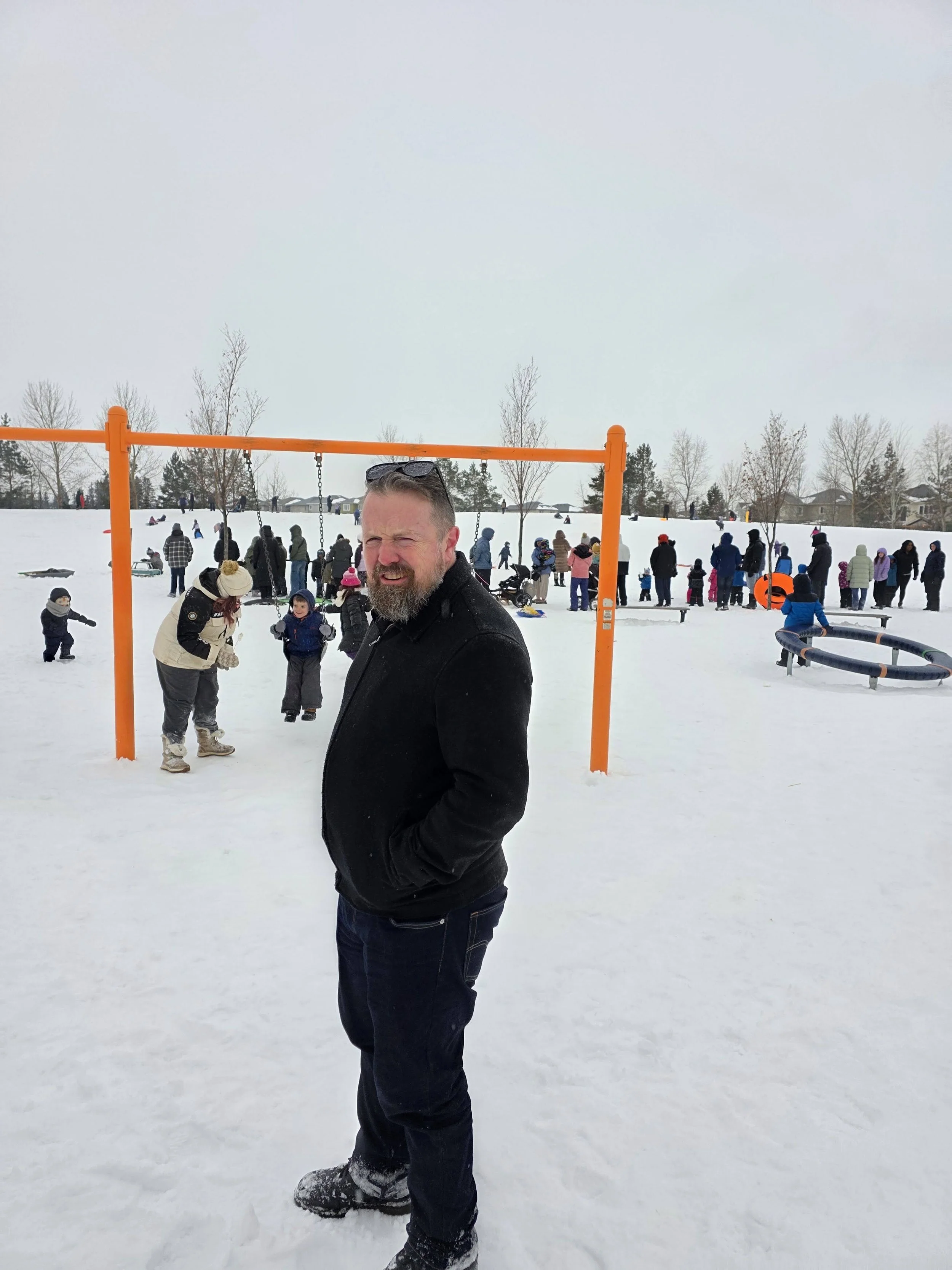 A man standing in the snow at a playground during winter, with children and adults in the background playing and socializing, and snow-covered trees and houses visible in the distance.